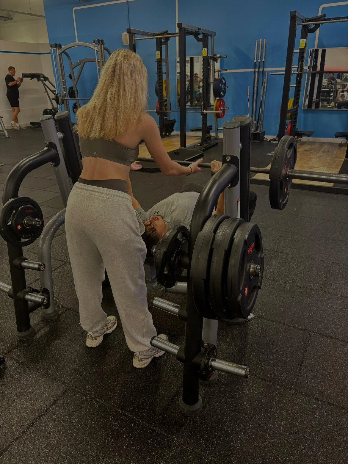 A woman spotting for a man who is lifting weights on a bench press at a gym.