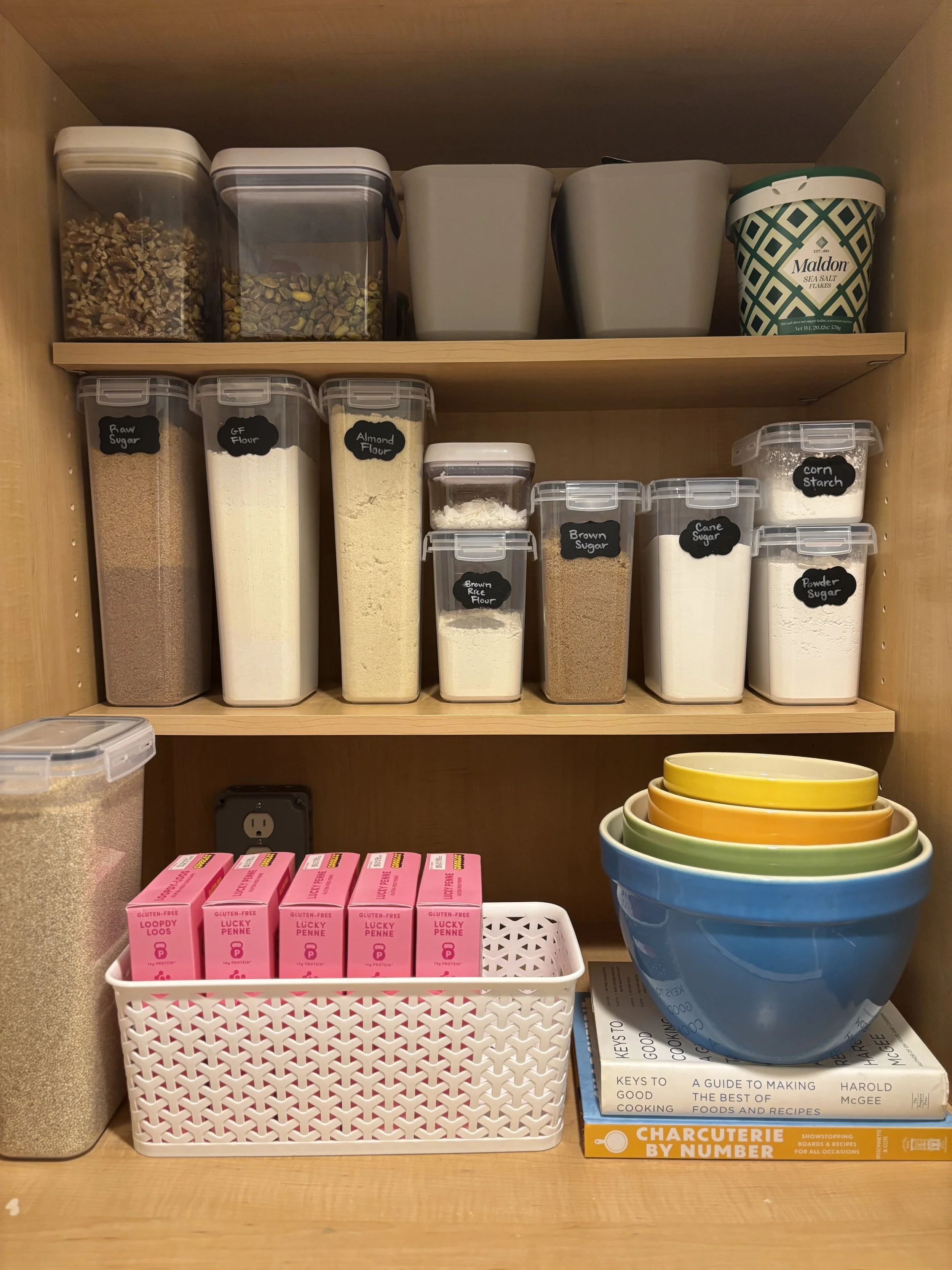 Pantry shelf with various containers holding dried goods, bowls stacked, and cookbooks.