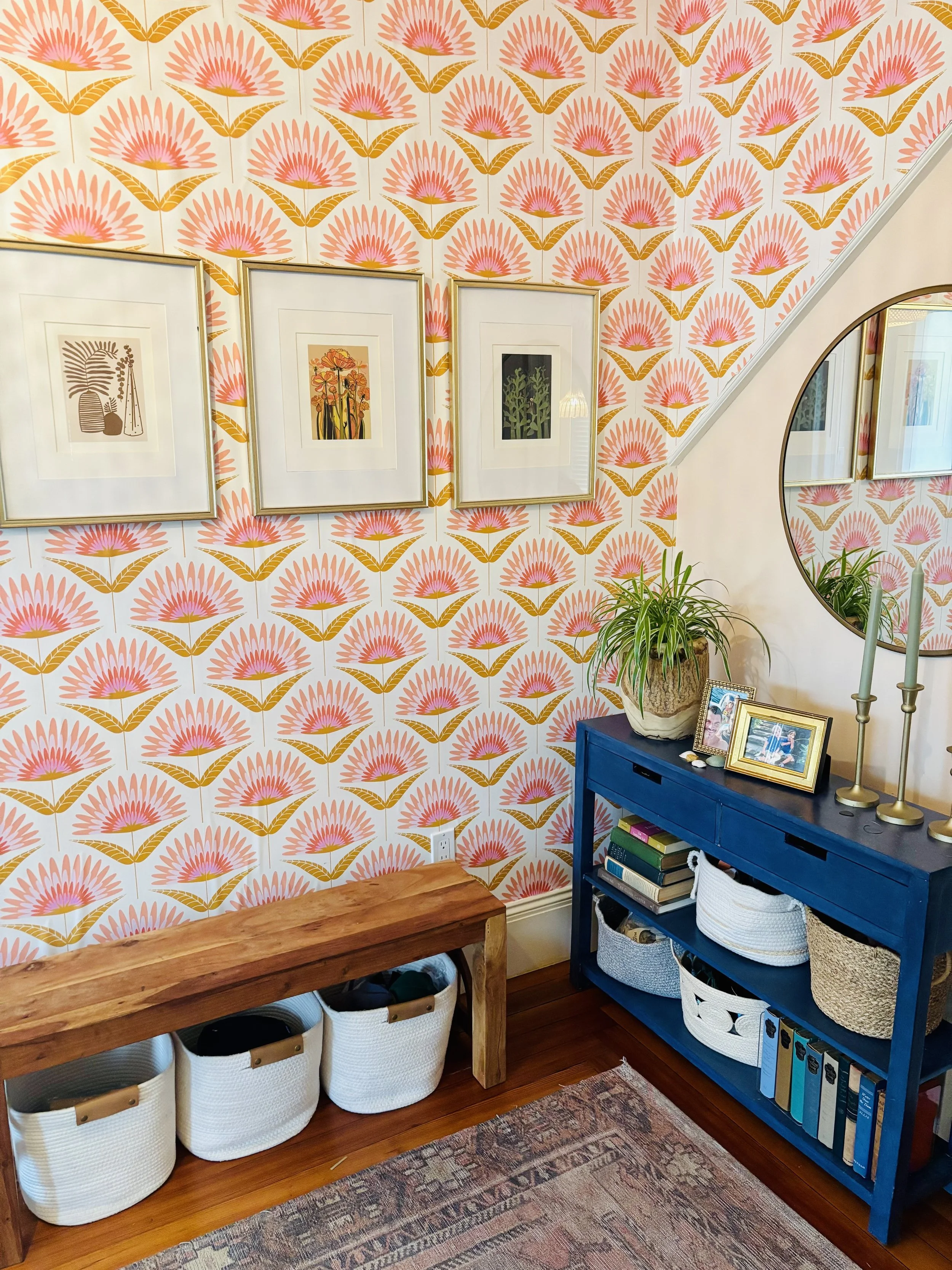 Decorative interior wall with framed artwork, a potted plant on a blue console table, photo frames, candlesticks, and baskets under a wooden bench, with patterned wallpaper and a mirror reflecting part of the room.