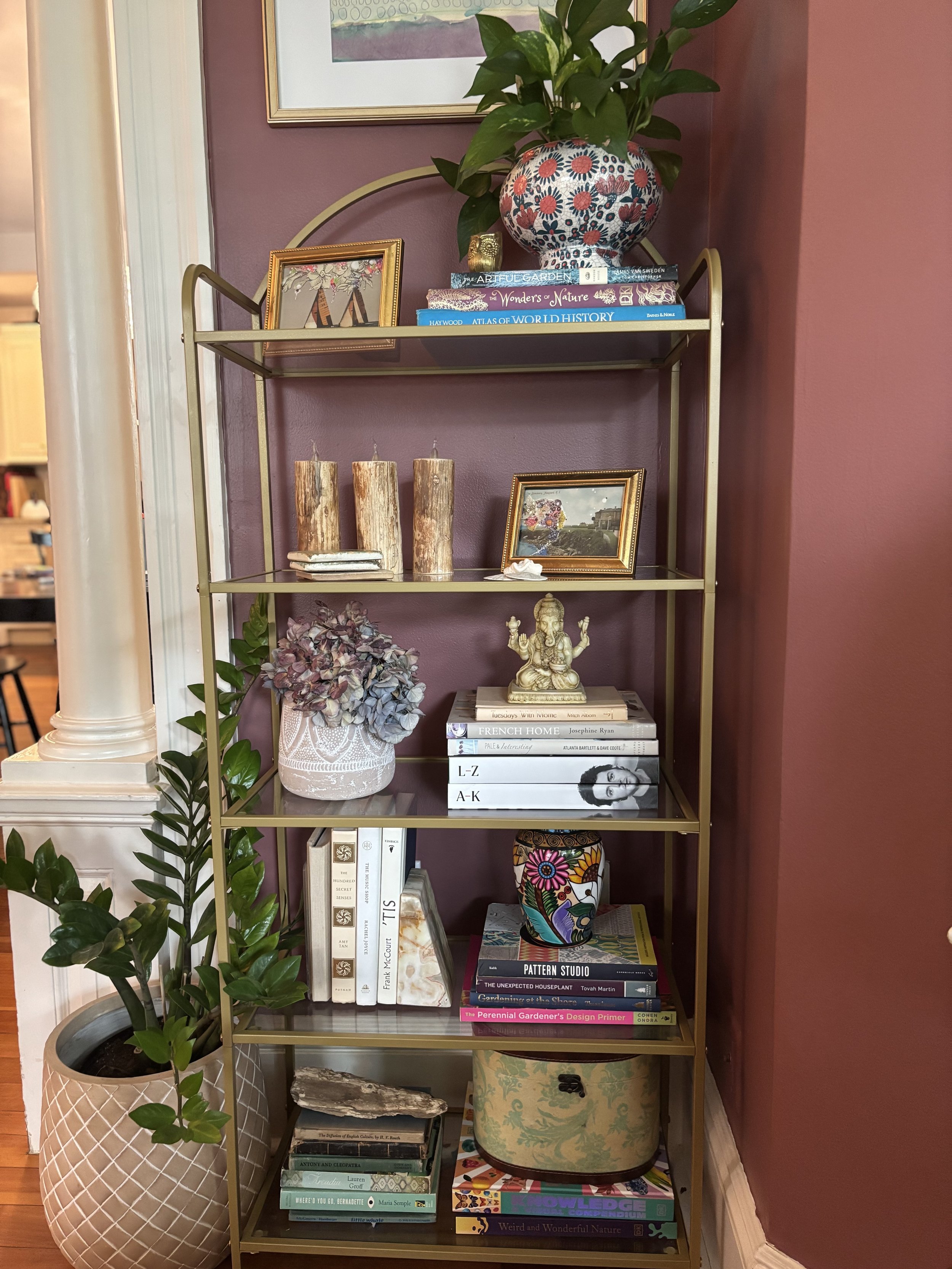 Gold-colored shelving unit with five shelves containing books, framed photos, a decorative vase with flowers, candles, a small statue, a potted plant, and decorative items against a burgundy wall.