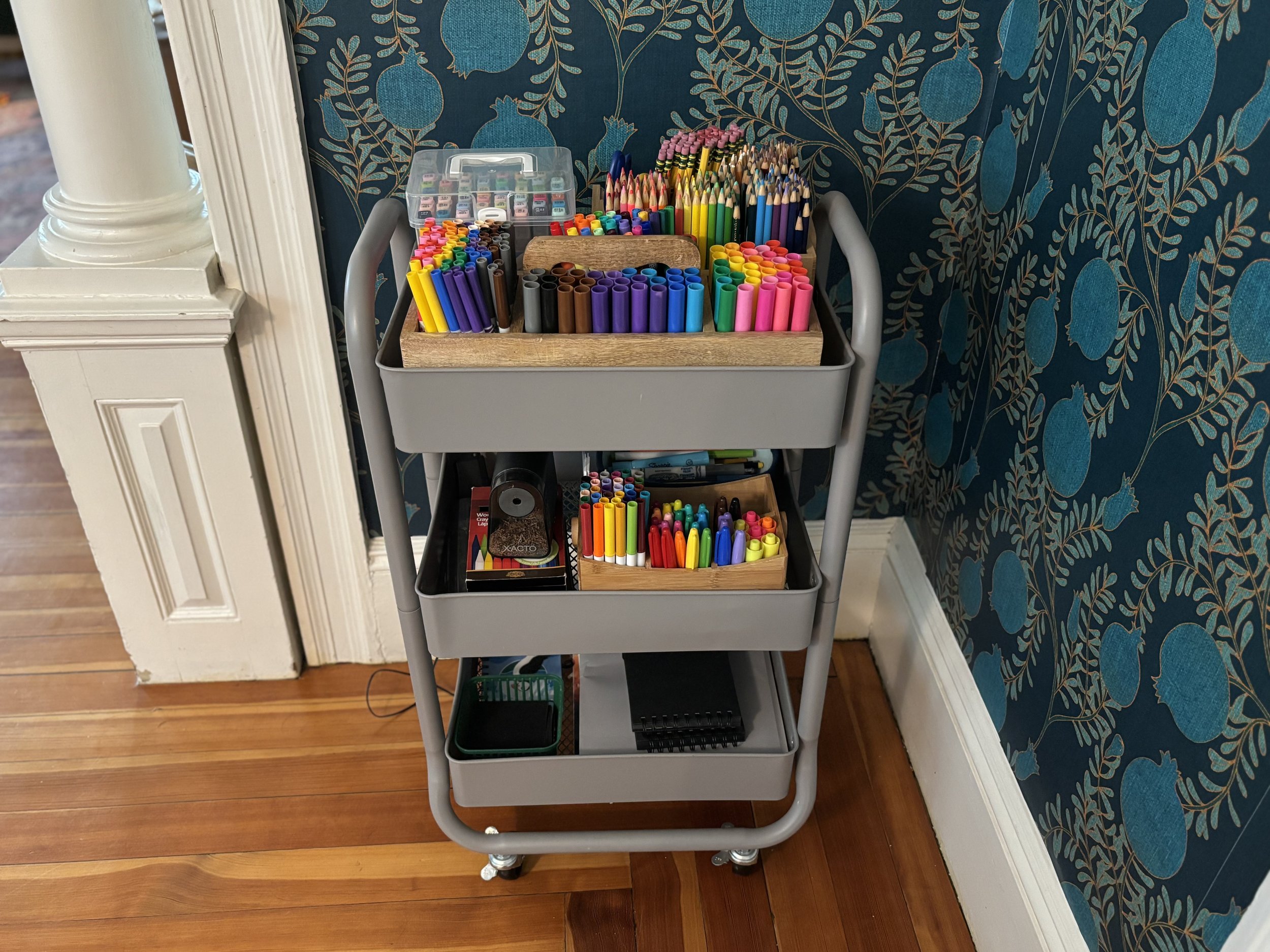 A gray multi-tiered cart filled with colorful writing and drawing supplies, including markers, colored pencils, and craft materials, positioned against a blue patterned wallpapered wall on a wooden floor.