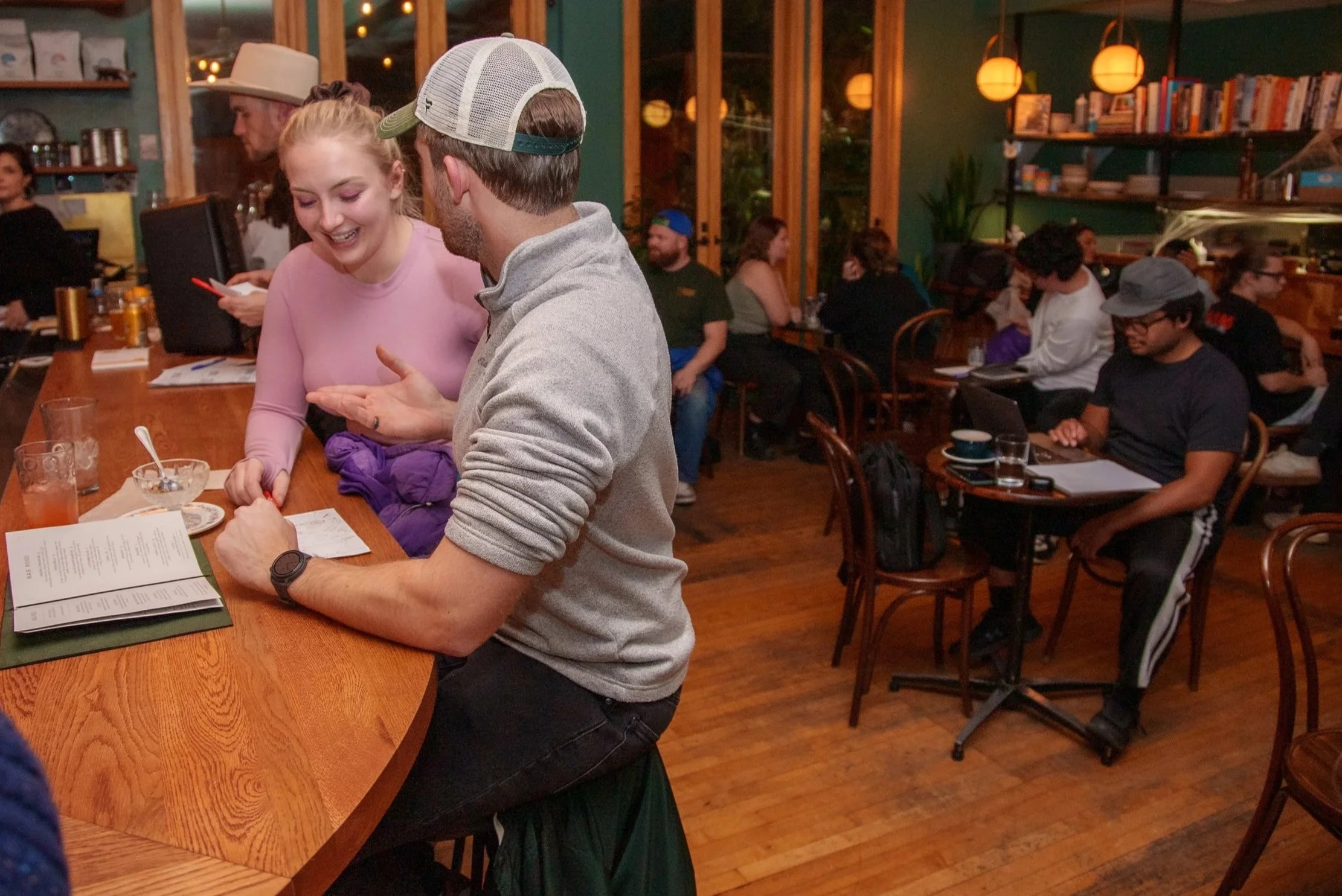 People dining in a cozy restaurant with warm lighting, wooden floors, and bookshelves on the wall, with two people engaged in conversation at the foreground area.