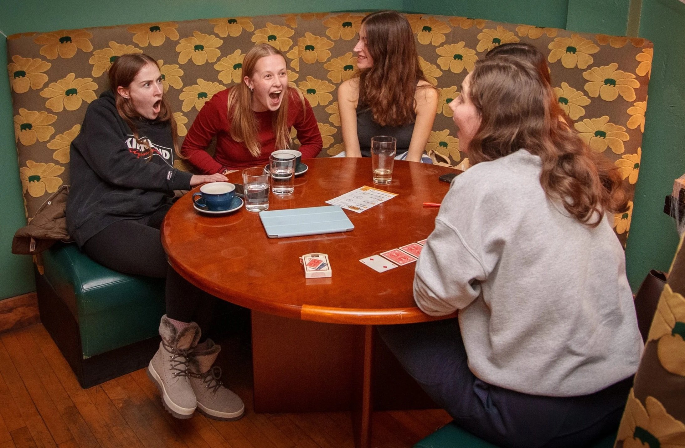Five women sitting at a round wooden table in a warmly lit restaurant decorated with floral-patterned wallpaper and artwork. They are engaged in a lively conversation, laughing, during a trivia game.