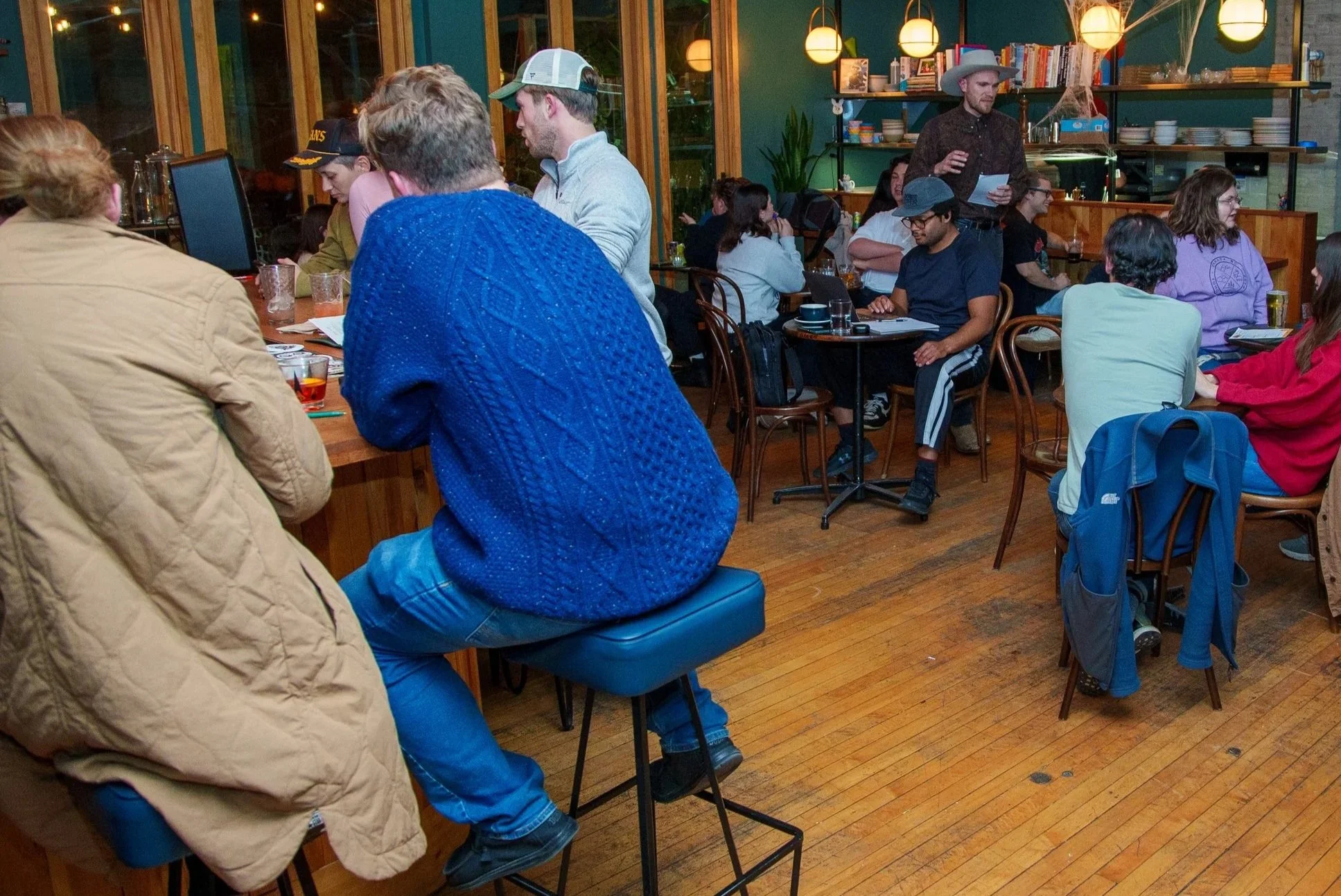 People gathered in a cozy, dimly lit cafe with green walls, wooden floors, and bookshelves.