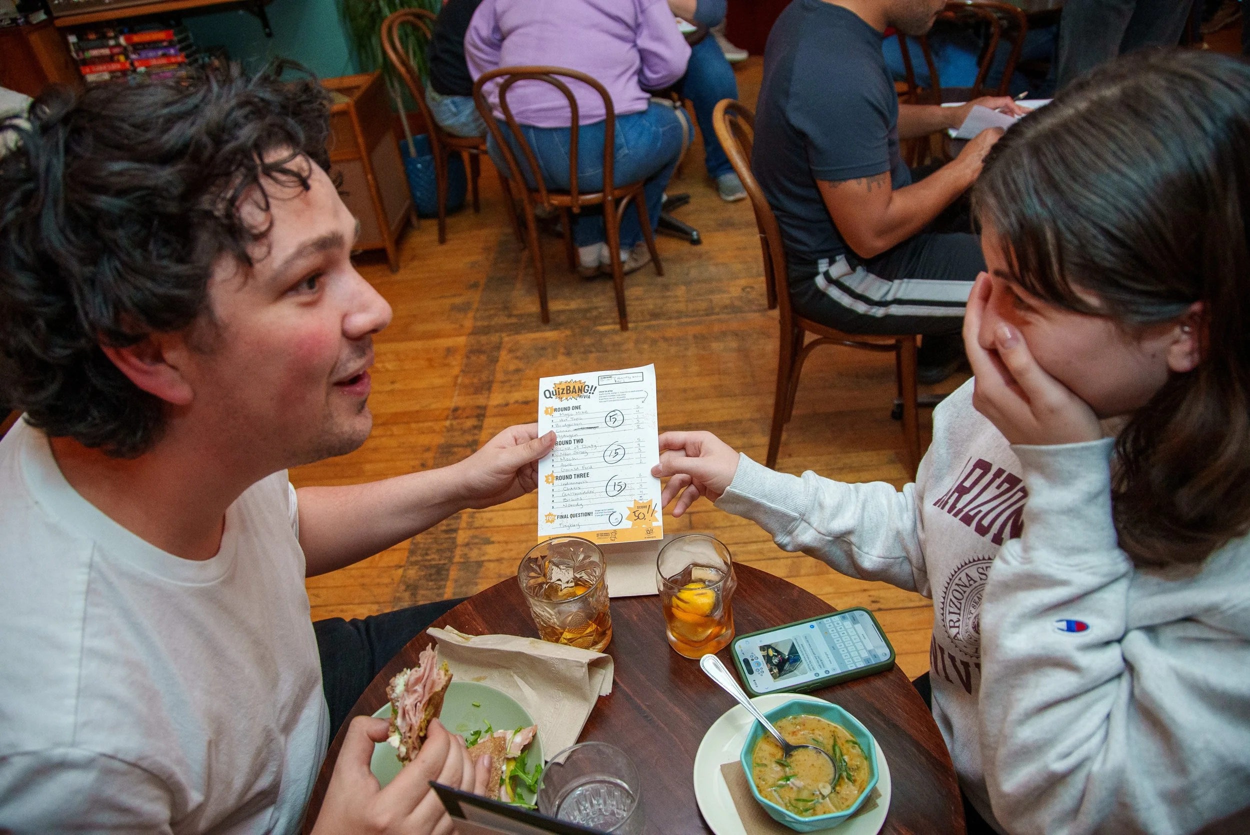 Two people eating at a small wooden table, laughing and sharing a QuizBang trivia answer sheet.
