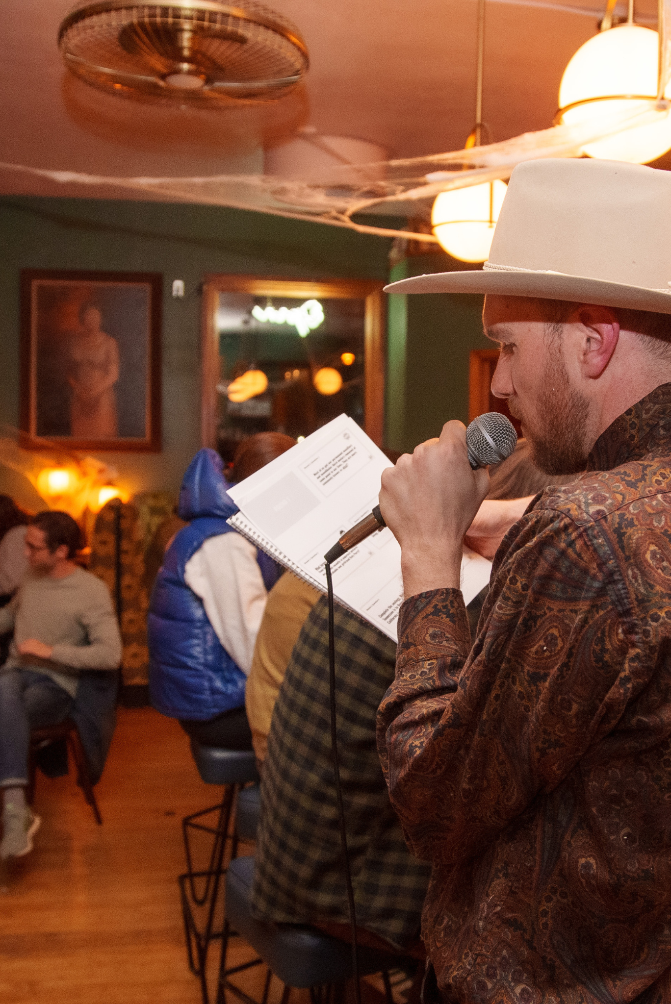 A man wearing a white cowboy hat and patterned shirt is speaking into a microphone while reading from a QuizBang trivia notebook in a warmly lit restaurant or bar with patrons in the background.
