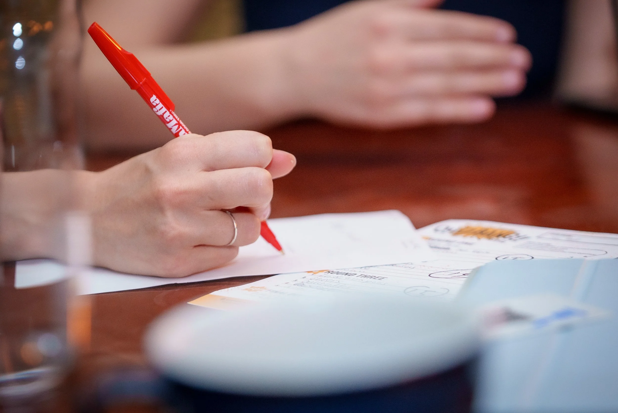 A person holding a red QuizBang trivia pen writing on paper at a wooden table.