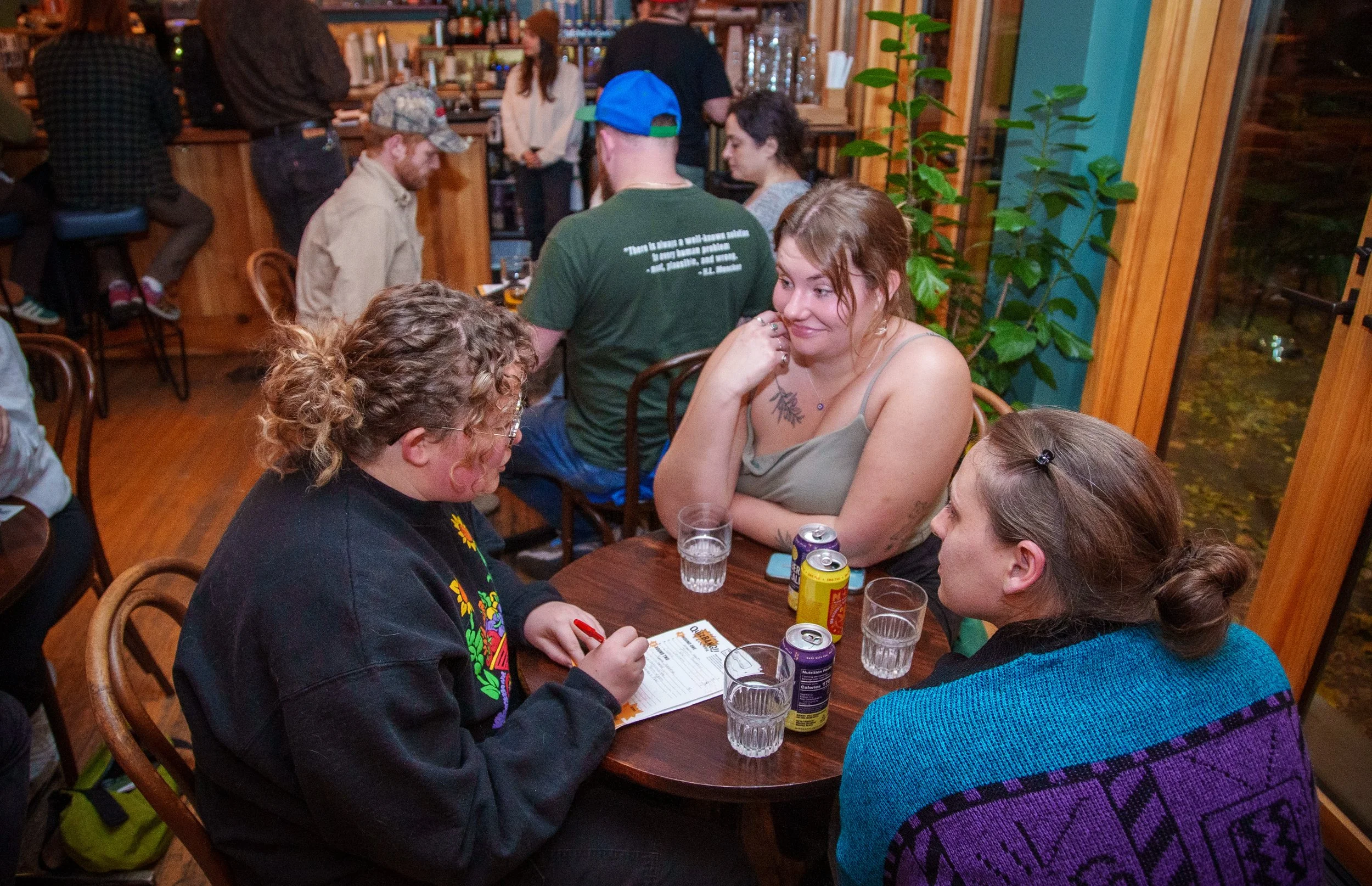 Three women are sitting at a table in a cozy restaurant, engaged in conversation. The table has four glasses of water, several canned drinks, and a menu. The woman on the left has curly hair, glasses, and is wearing a black sweatshirt with colorful e