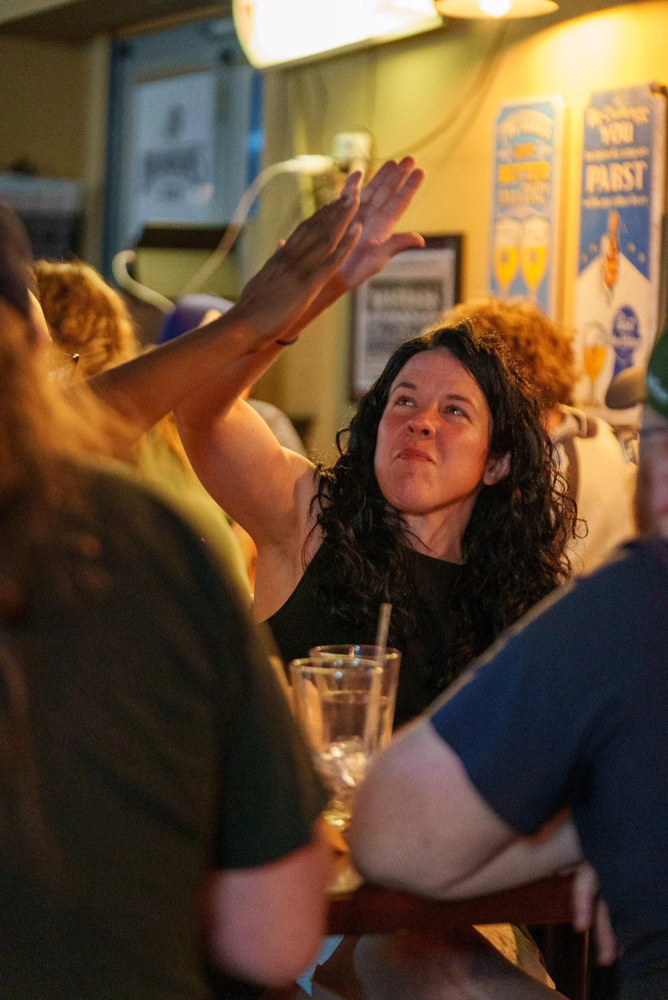 A woman with curly dark hair and a black sleeveless top is giving a high five to another person in a crowded restaurant.