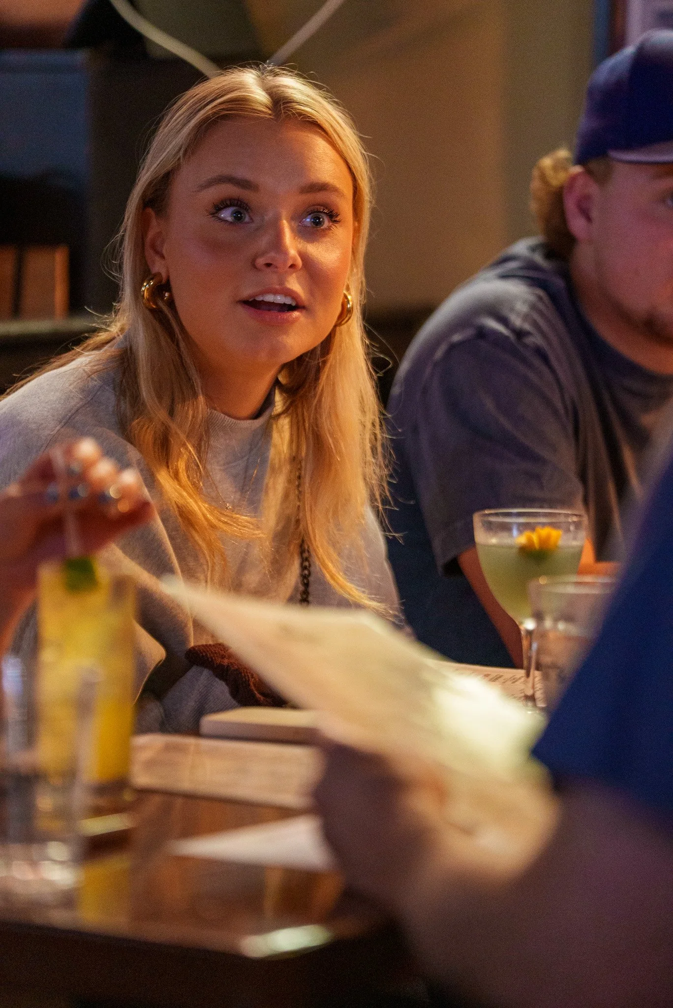 A young woman with blonde hair, looking surprised as she talks to her friends during a trivia game.