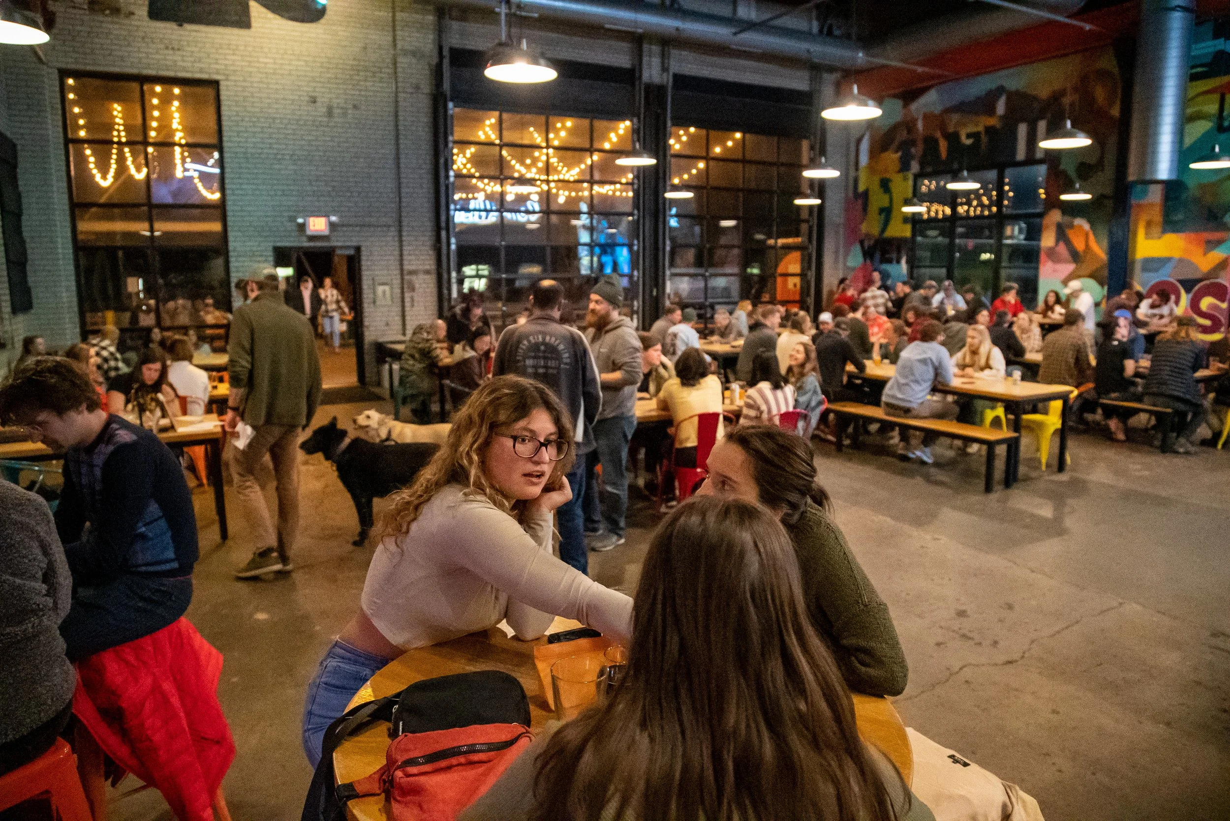 People gathered inside a lively bar with colorful murals, string lights, and a large crowd seated at long tables, engaging in conversations.