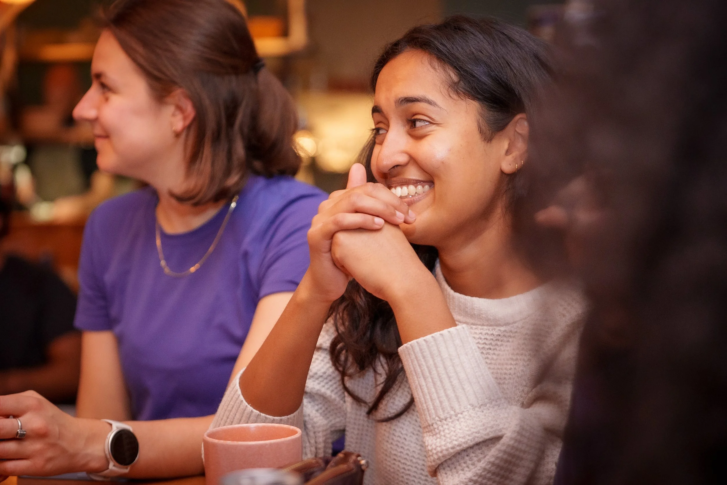 A woman with dark hair and a light pink sweater smiling and resting her chin on her clasped hands while playing trivia.