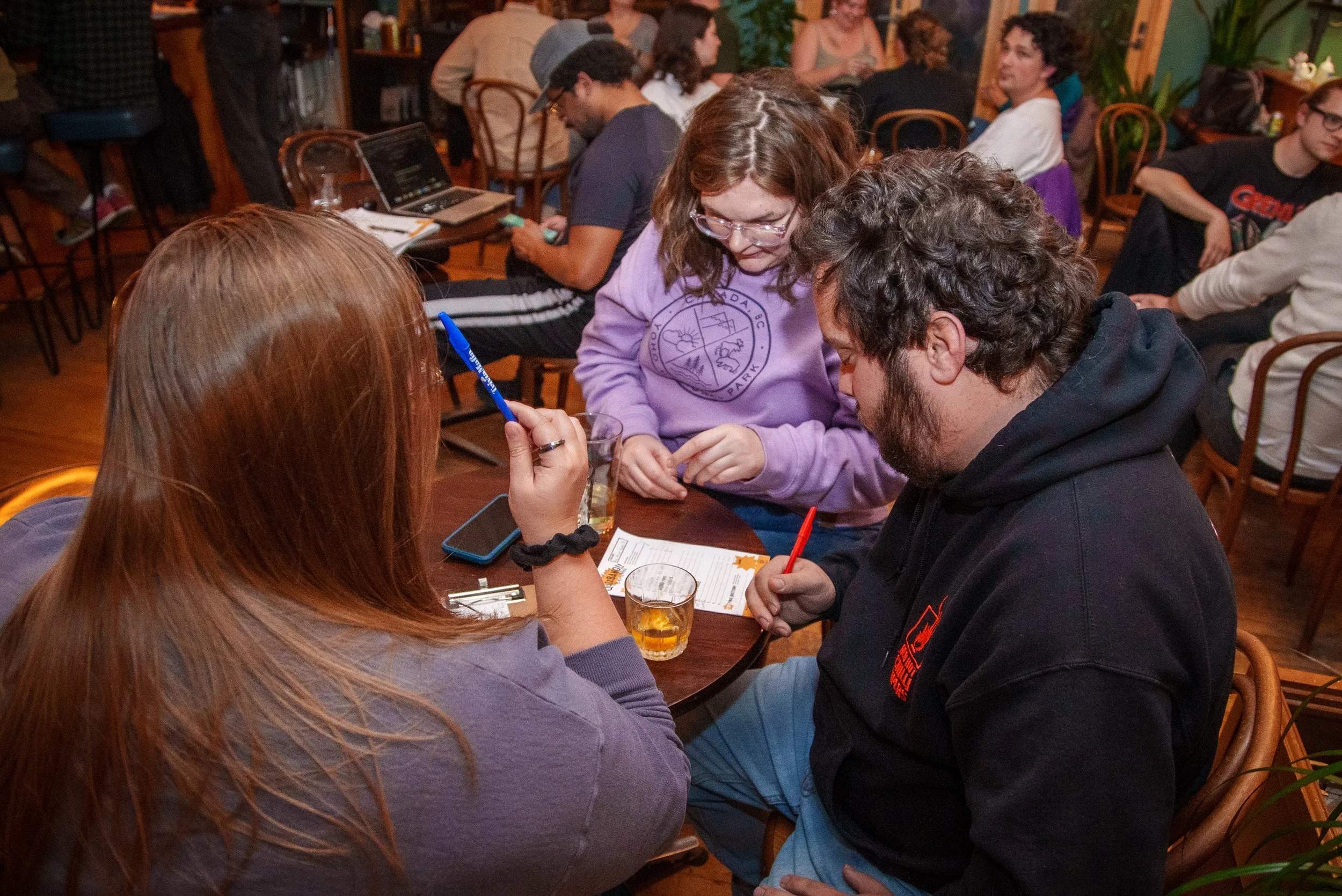 Three people sitting at a table in a lively restaurant, engaged in a game or activity, with drinks and phone on the table. The background shows other patrons and a warm, decorated interior.