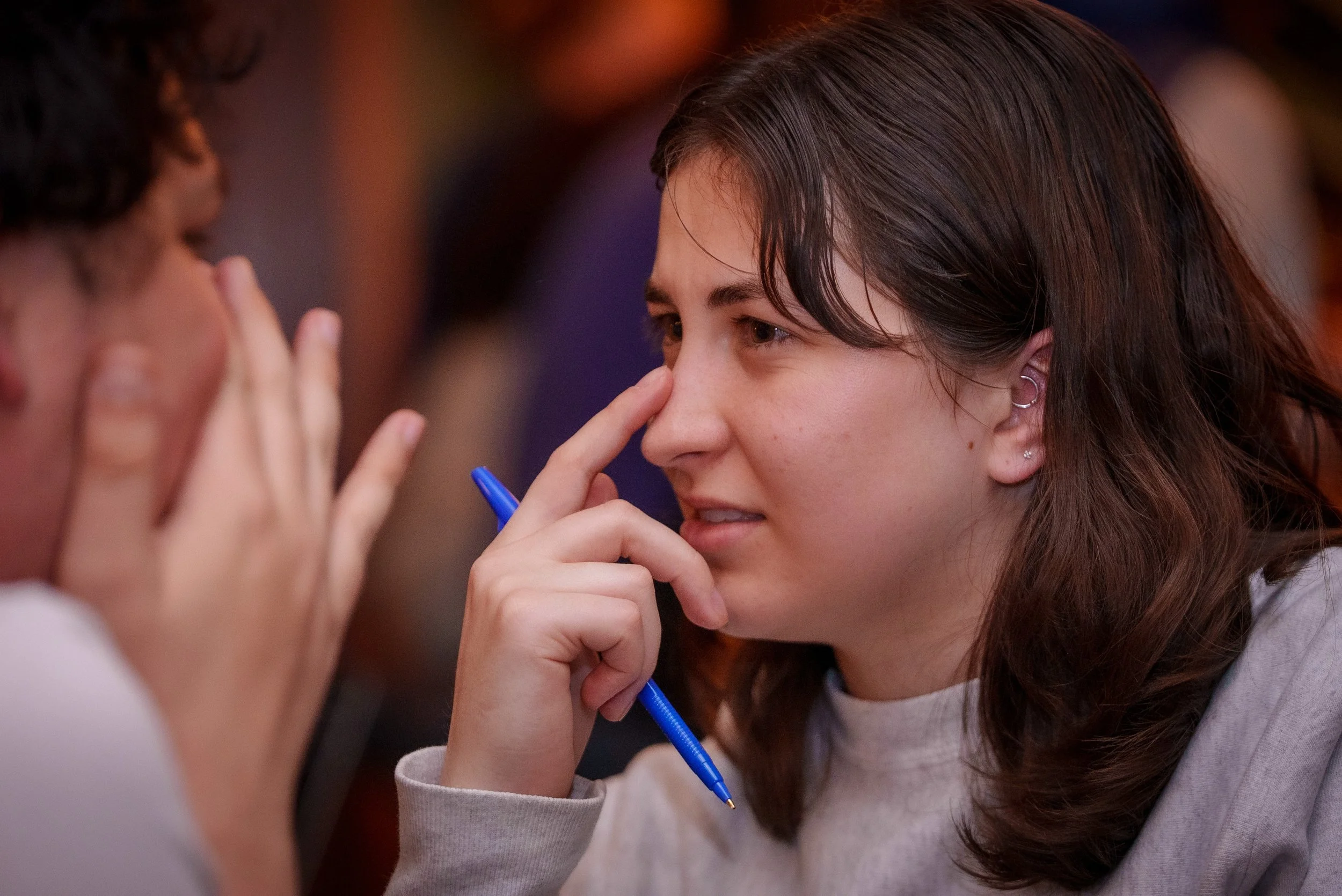 A young woman with brown hair, holding a blue pen in her hand, with one finger on her nose while she thinks. 