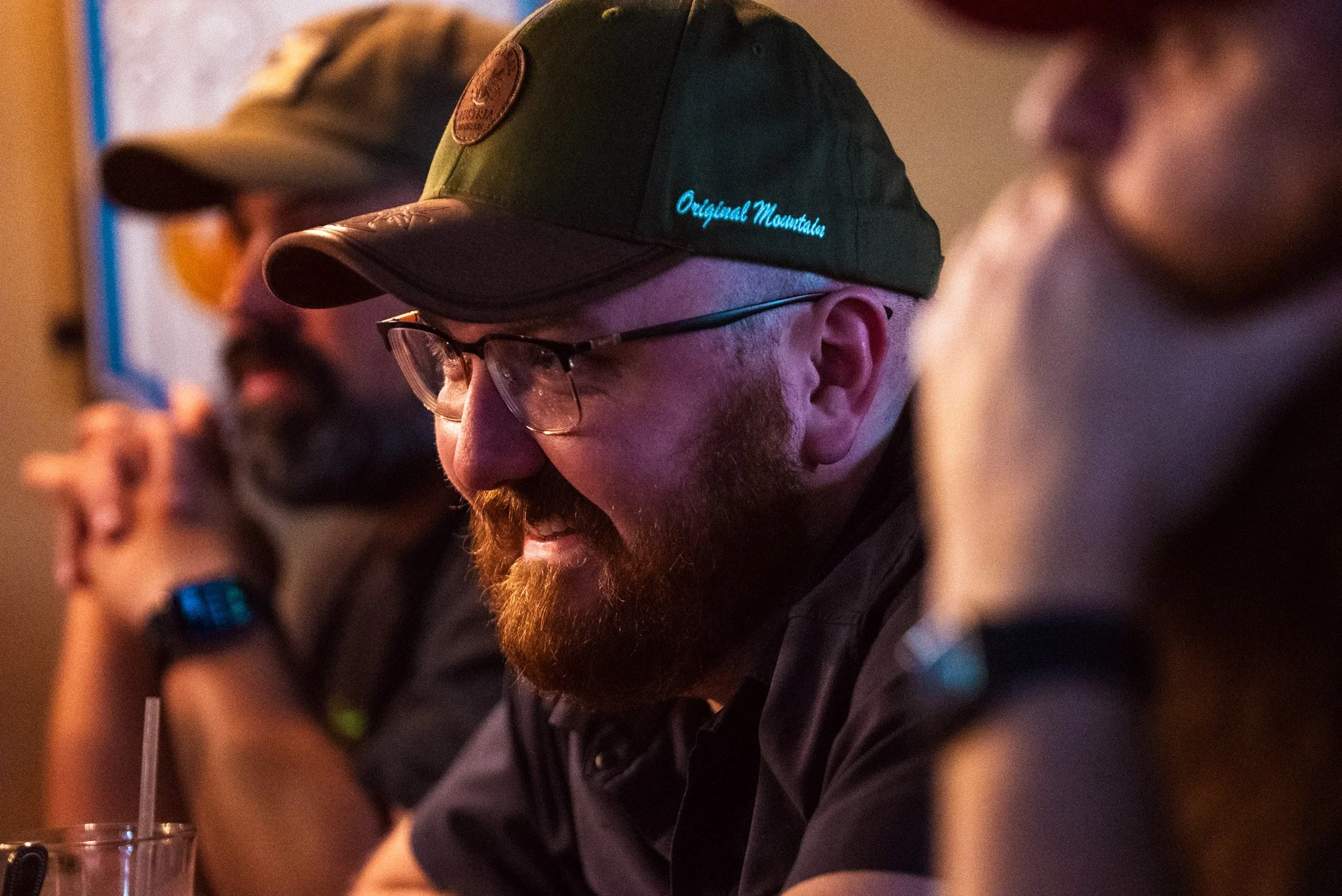 A man with a beard, glasses, and a green baseball cap with the words 'Original Mountain' embroidered in blue. He is smiling and sitting at a table with a drink and straw. There are other people around him, partly visible, also at the table.