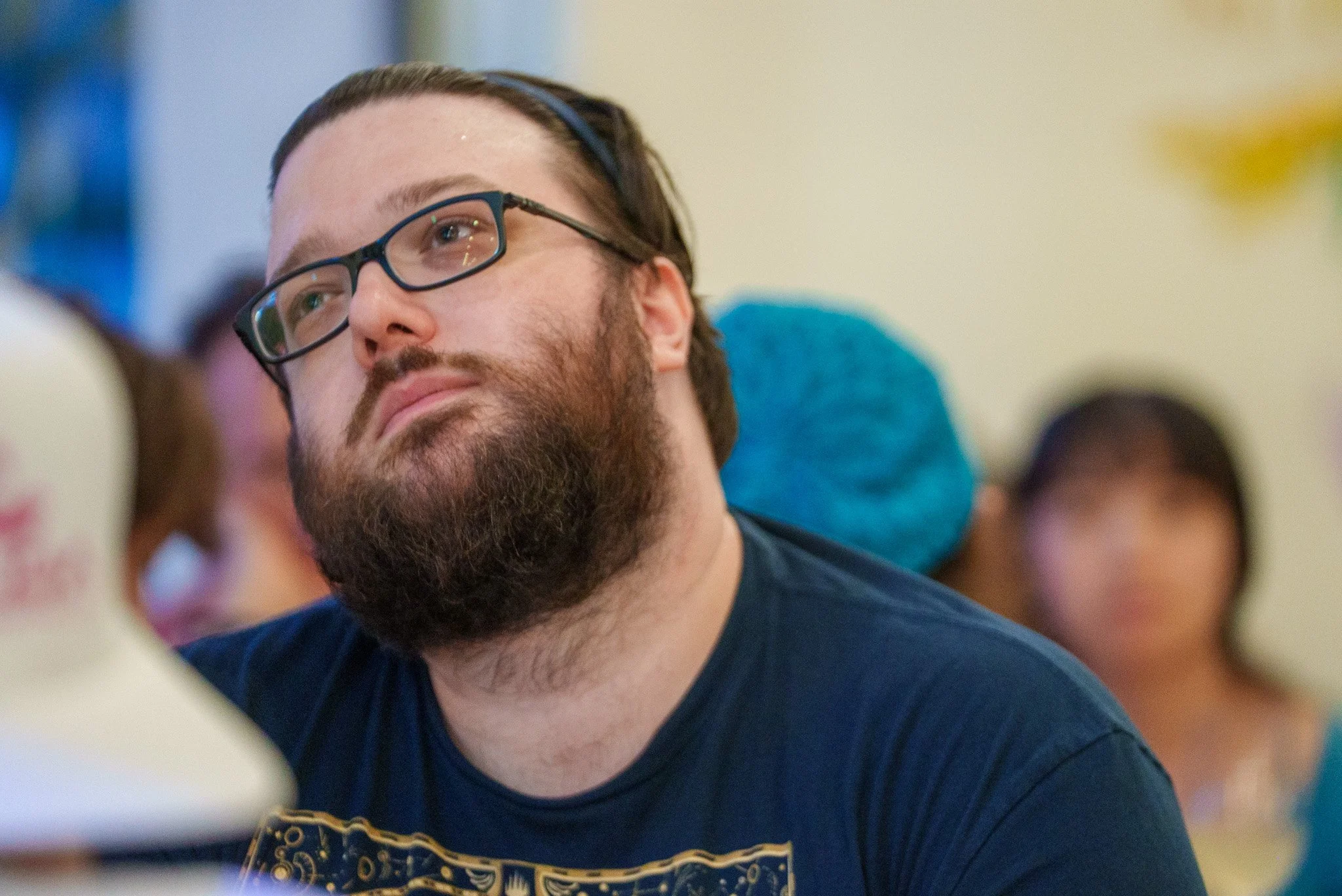 Man with glasses and a beard, looking thoughtful while trying to think of a trivia answer, with people in the background.