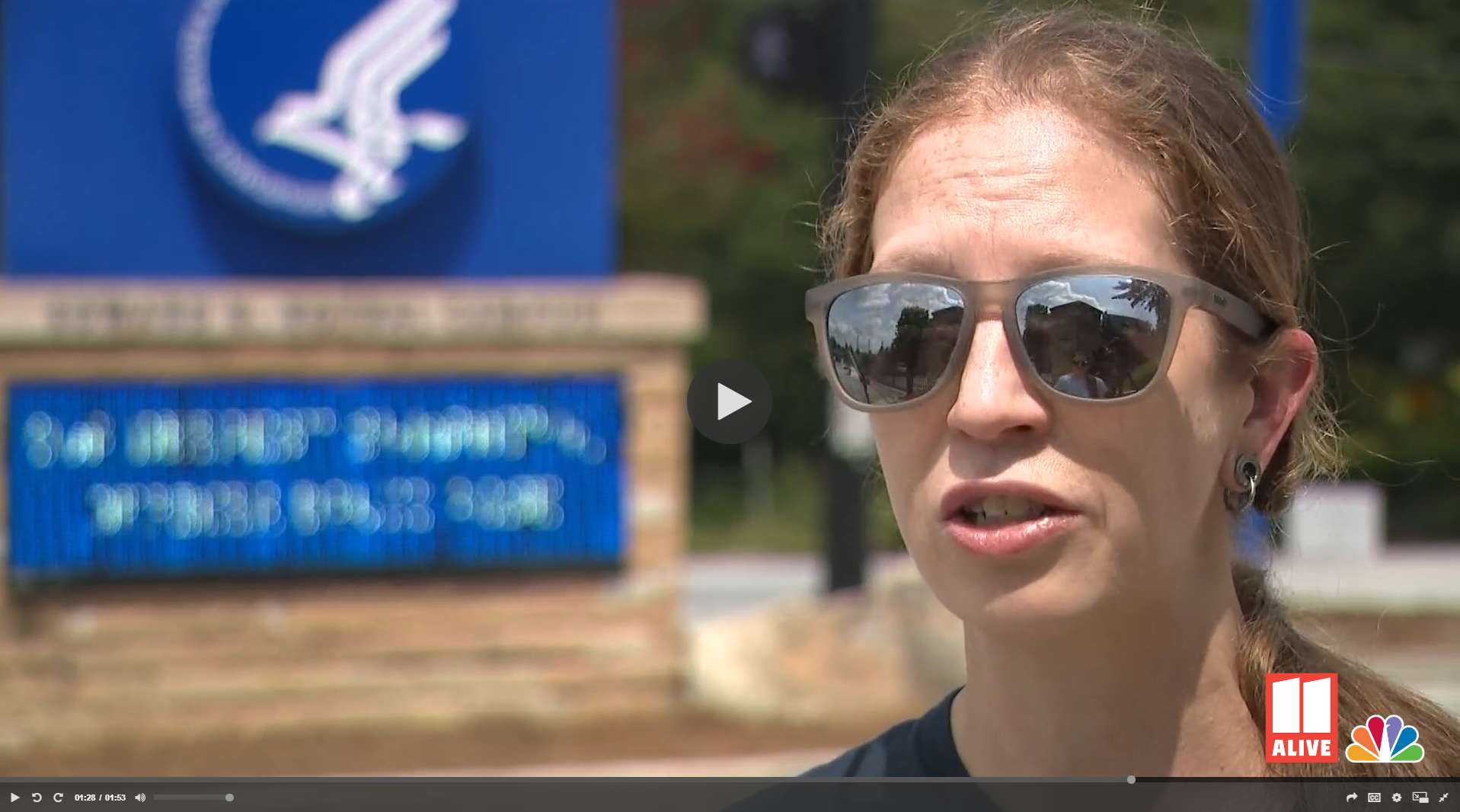 A woman wearing sunglasses is speaking outdoors in front of a sign with the NBC logo and a blue bulletin board.
