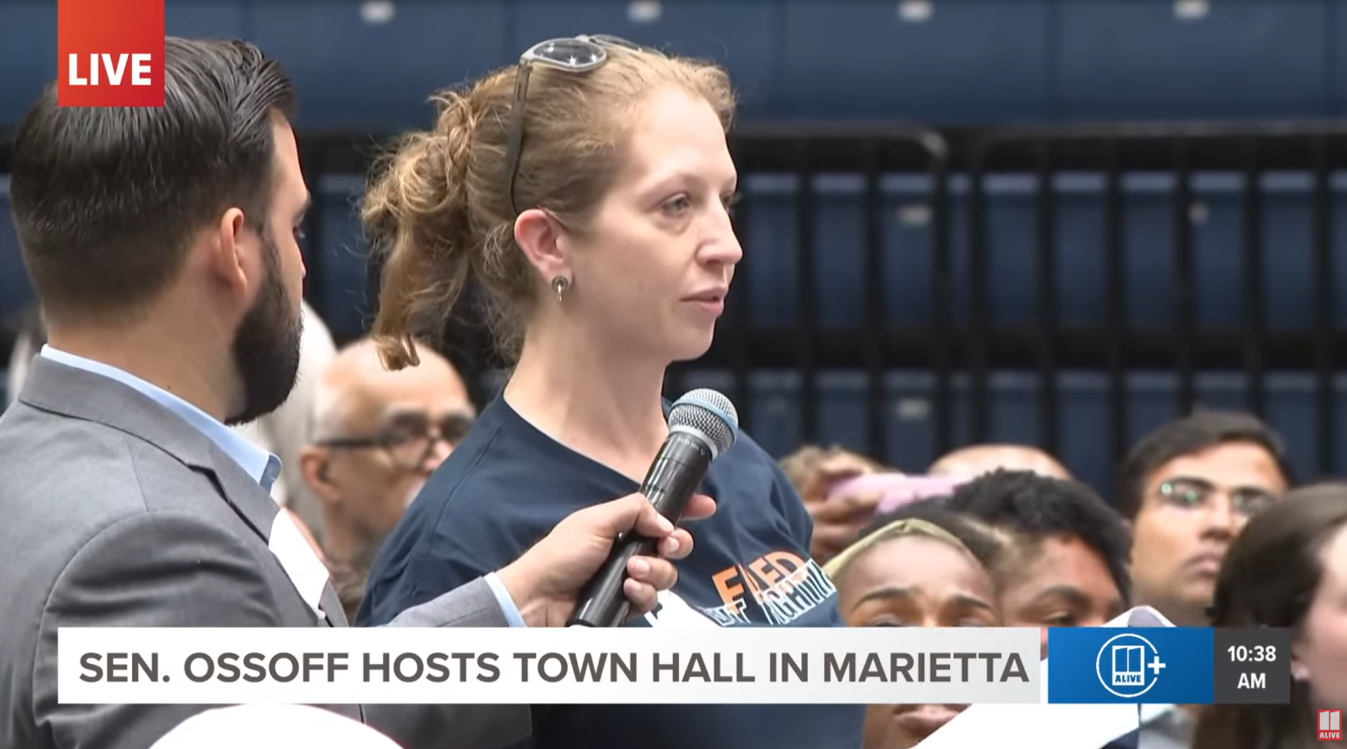 Senator Ossoff holding a microphone while speaking during a town hall in Marietta, with an audience and a news banner at the bottom.
