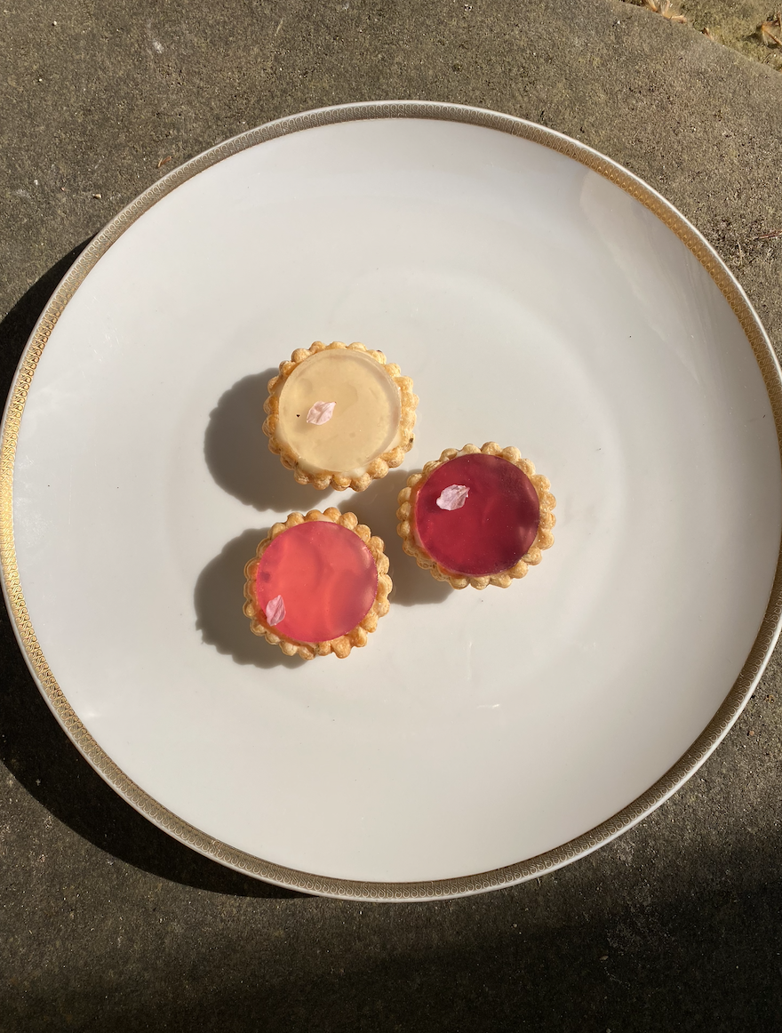 Trois tartelettes aux couleurs pastel sur une assiette blanche, décorées avec de petites fleurs de cerisier en papier. Vue de dessus, en plein soleil, posées sur une surface en béton.