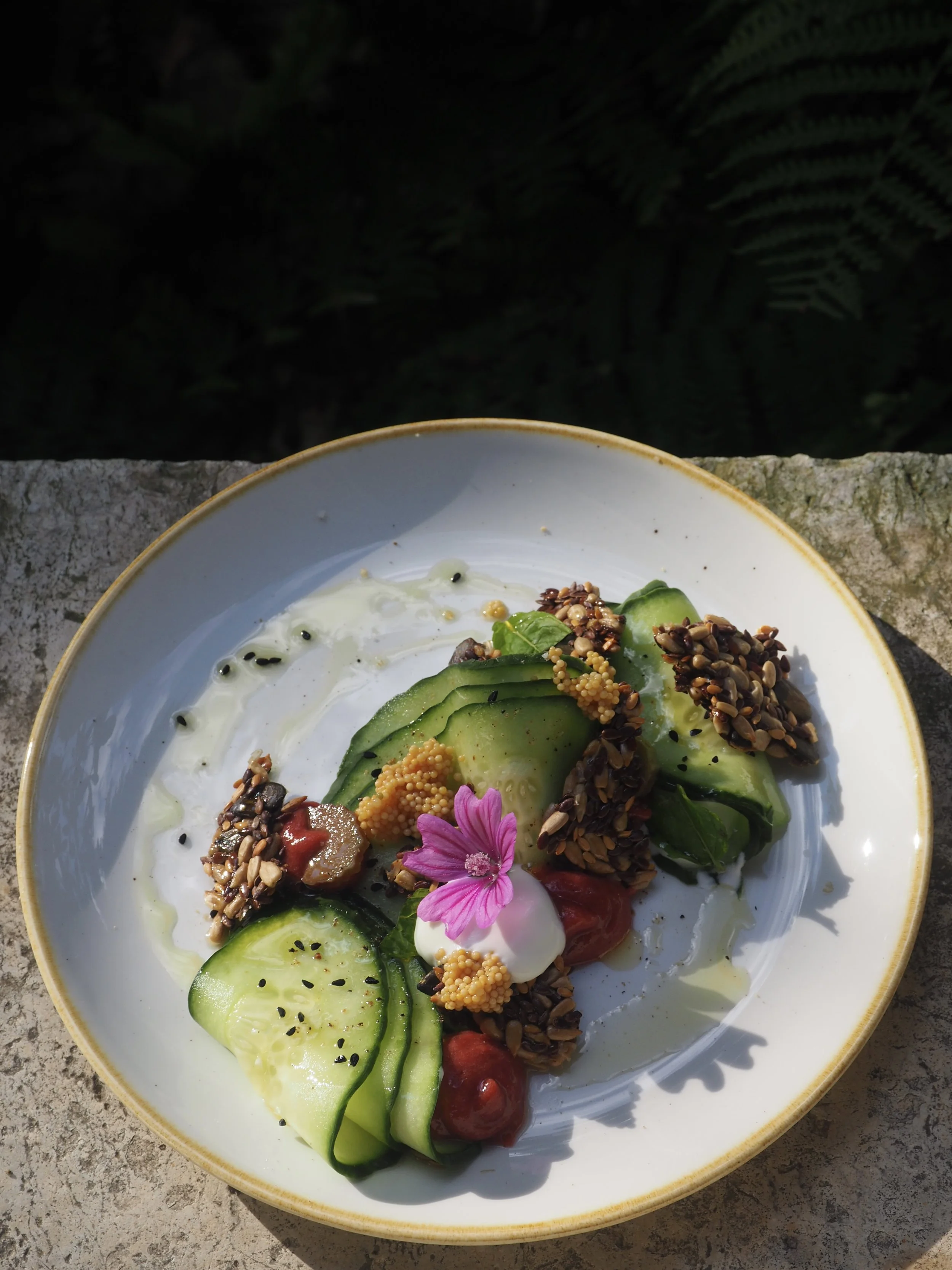 Salade de légumes avec concombre, tomates, œufs de caille, graines, fleur comestible et sauce dans un bol blanc.