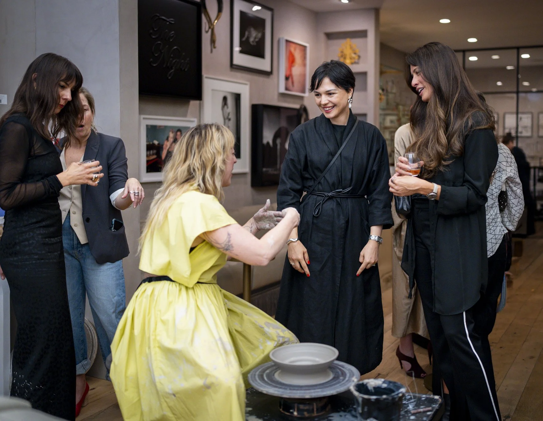 A group of five women, including one seated woman in a yellow dress with blonde hair, interacting and laughing at a social gathering in an art gallery or cafe setting. Four women are standing around her, holding drinks and engaging in conversation.