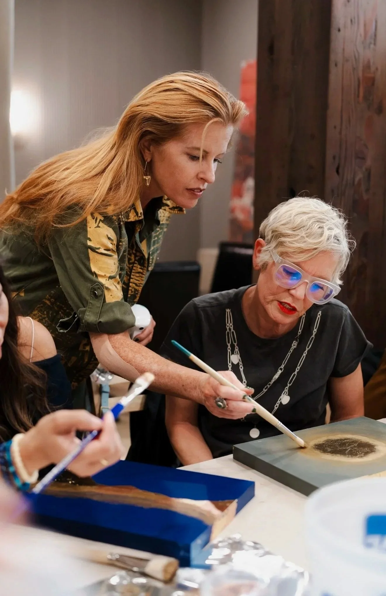 A woman with long red hair and a woman with short blonde hair and glasses are painting on a round canvas together while surrounded by others with art supplies.