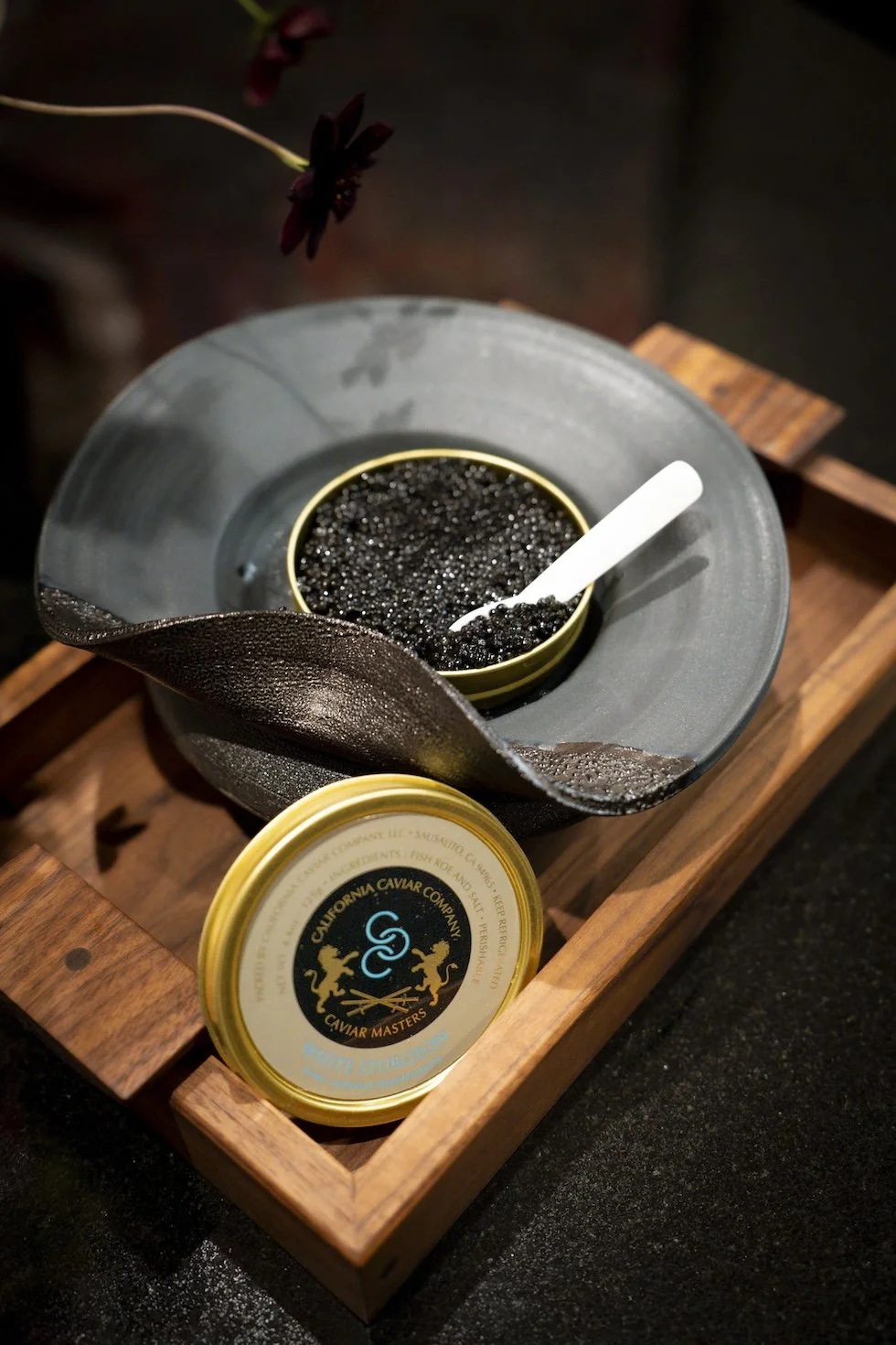 A container of black caviar with a spoon in a dish, placed inside a wooden tray with a label of California Caviar Company.