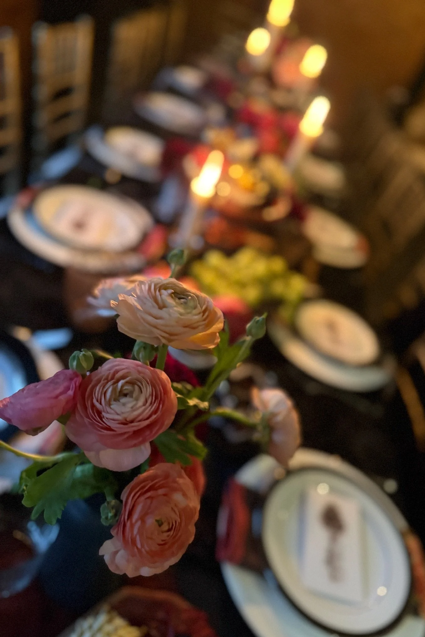 Bouquet of pink and orange ranunculus flowers in focus with a blurred background of various dishes, candles, and a dark table setting.