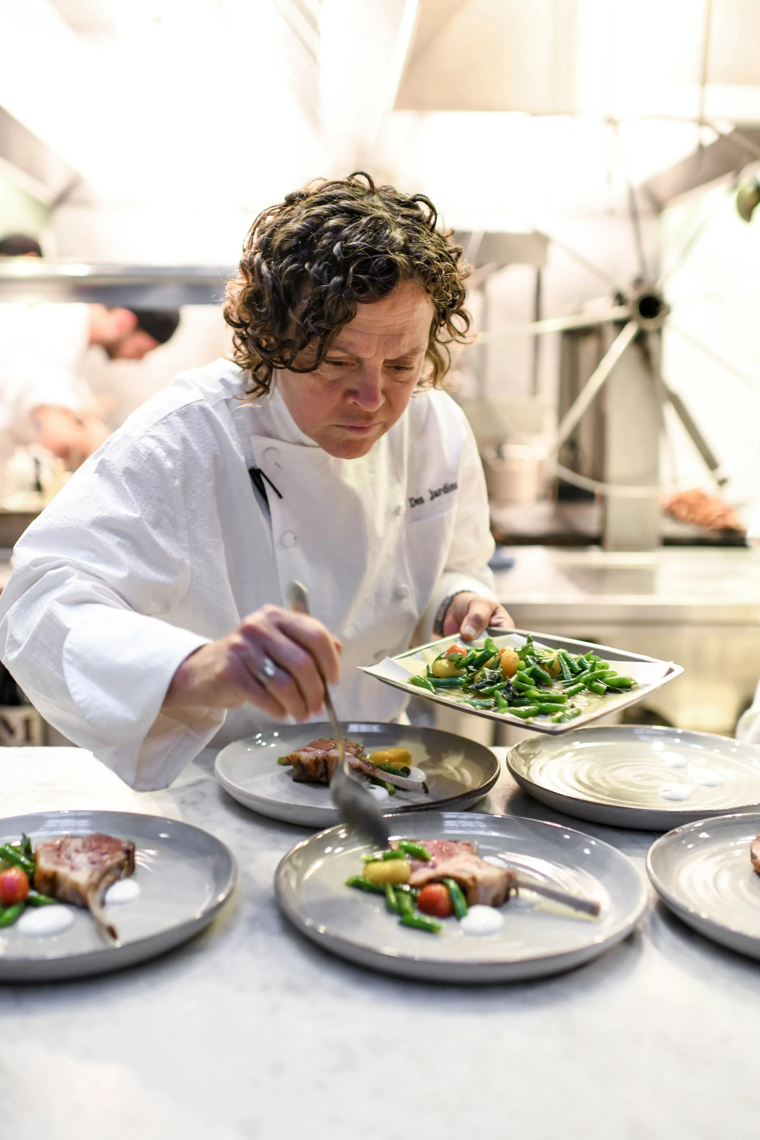 Chef plating a dish with green beans, cherry tomatoes, and potatoes, in a professional kitchen with other chefs in the background.