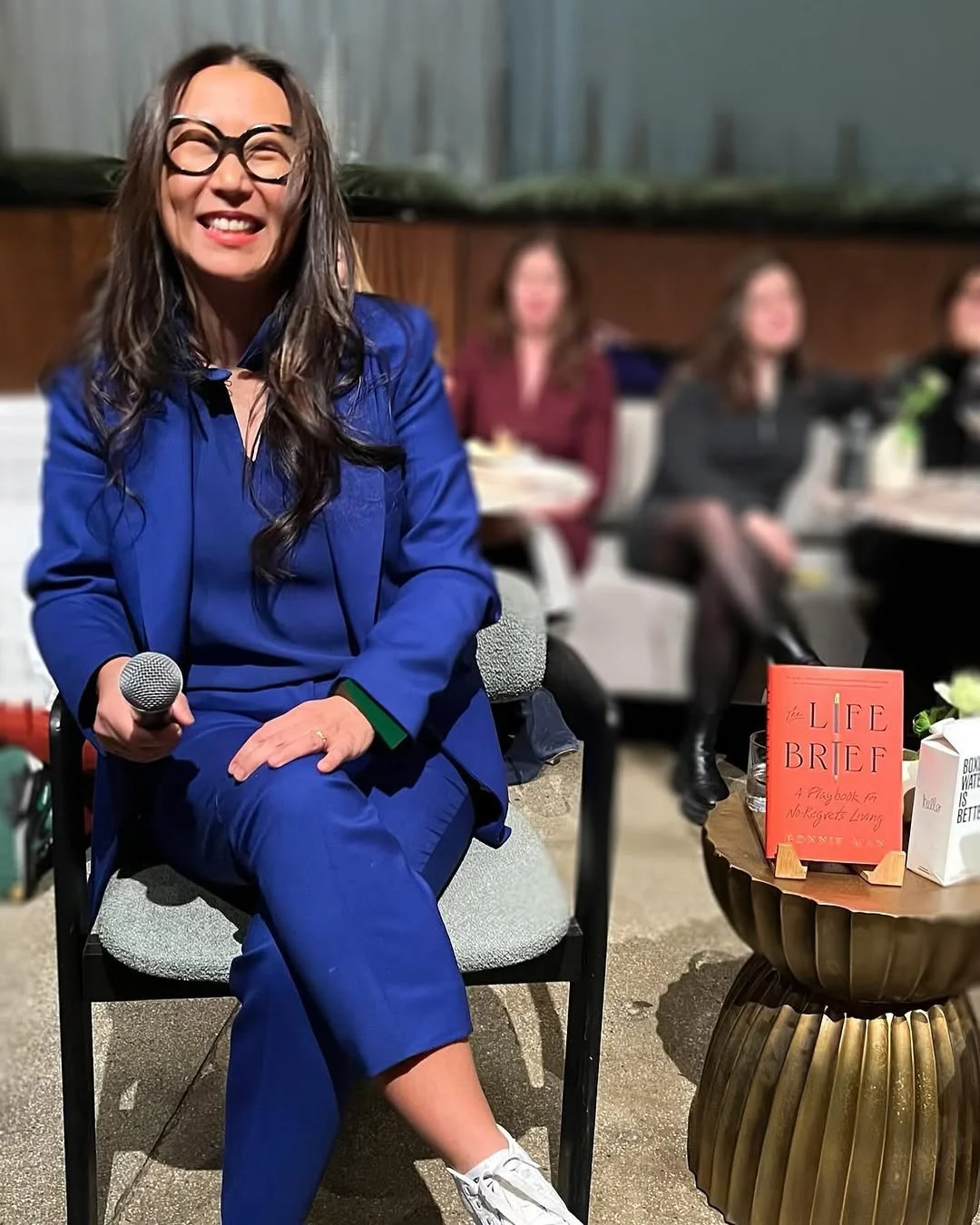 A woman in a blue outfit and glasses sitting on a chair, holding a microphone, smiling, with a group of women sitting behind her at a table.