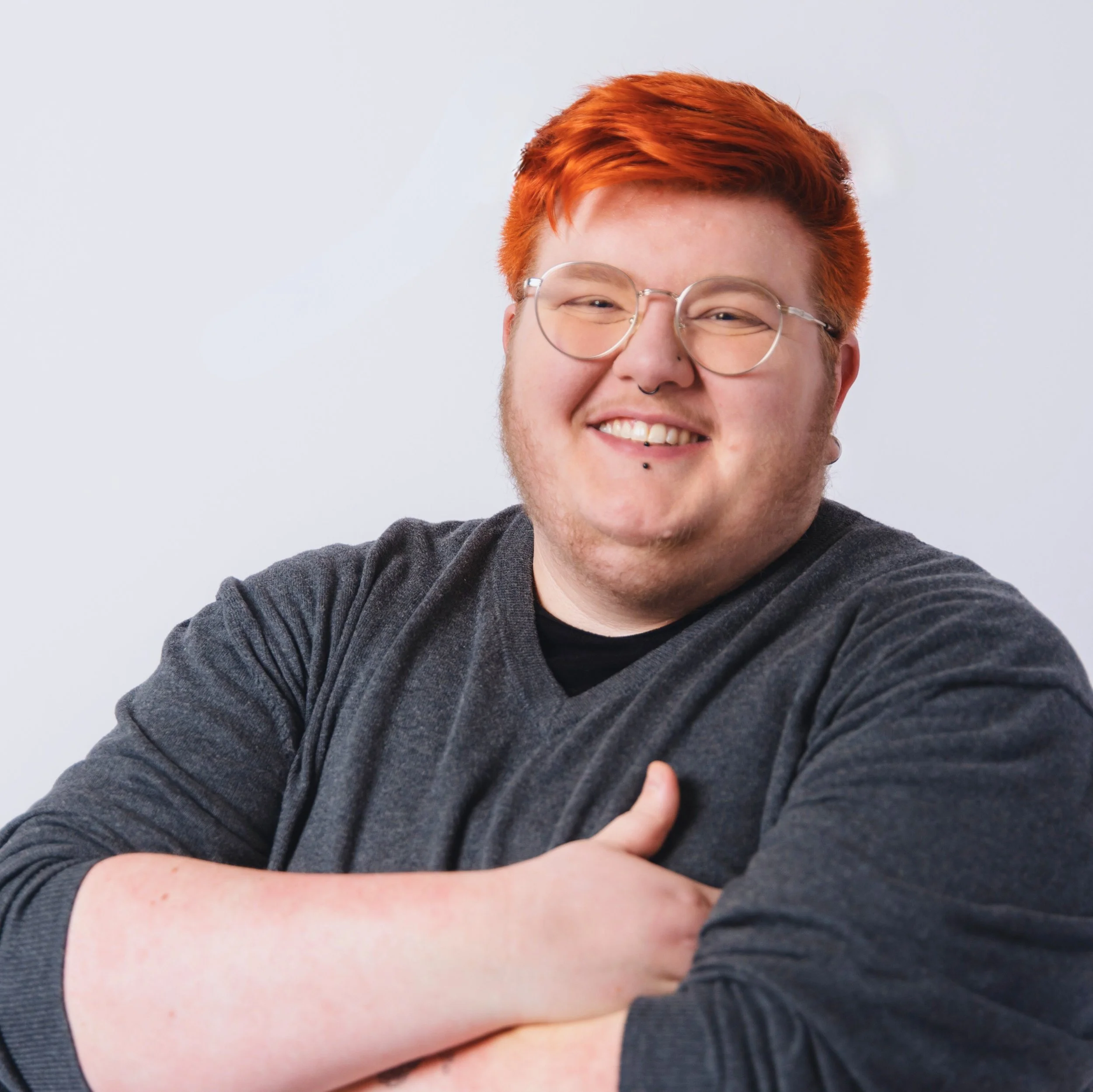 Image of Charley, they have a short dyed orange undercut and are wearing a grey sweater with their arms crossed over their chest. They are smiling and have a black lip piercing and gold glasses.
