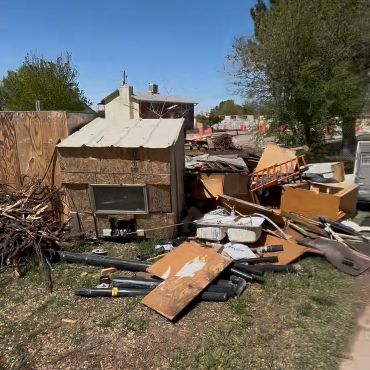Debris and destruction from a collapsed small wooden structure, scattered tools, wood, pipes, and trash on grass, with trees and a building in the background under a clear blue sky.