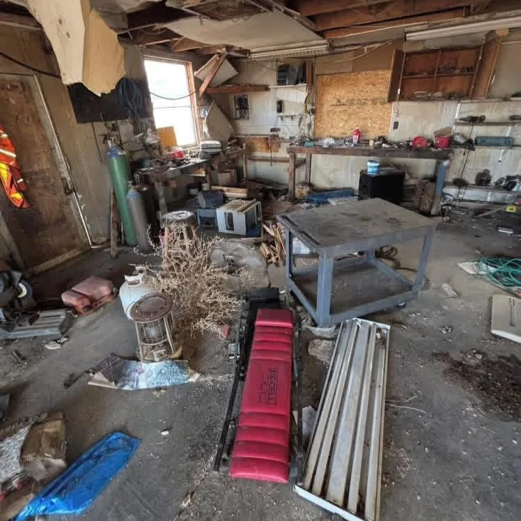 Disorganized garage with tools, furniture, and miscellaneous items scattered across the floor, exposed wooden beams, and a small window letting in light.