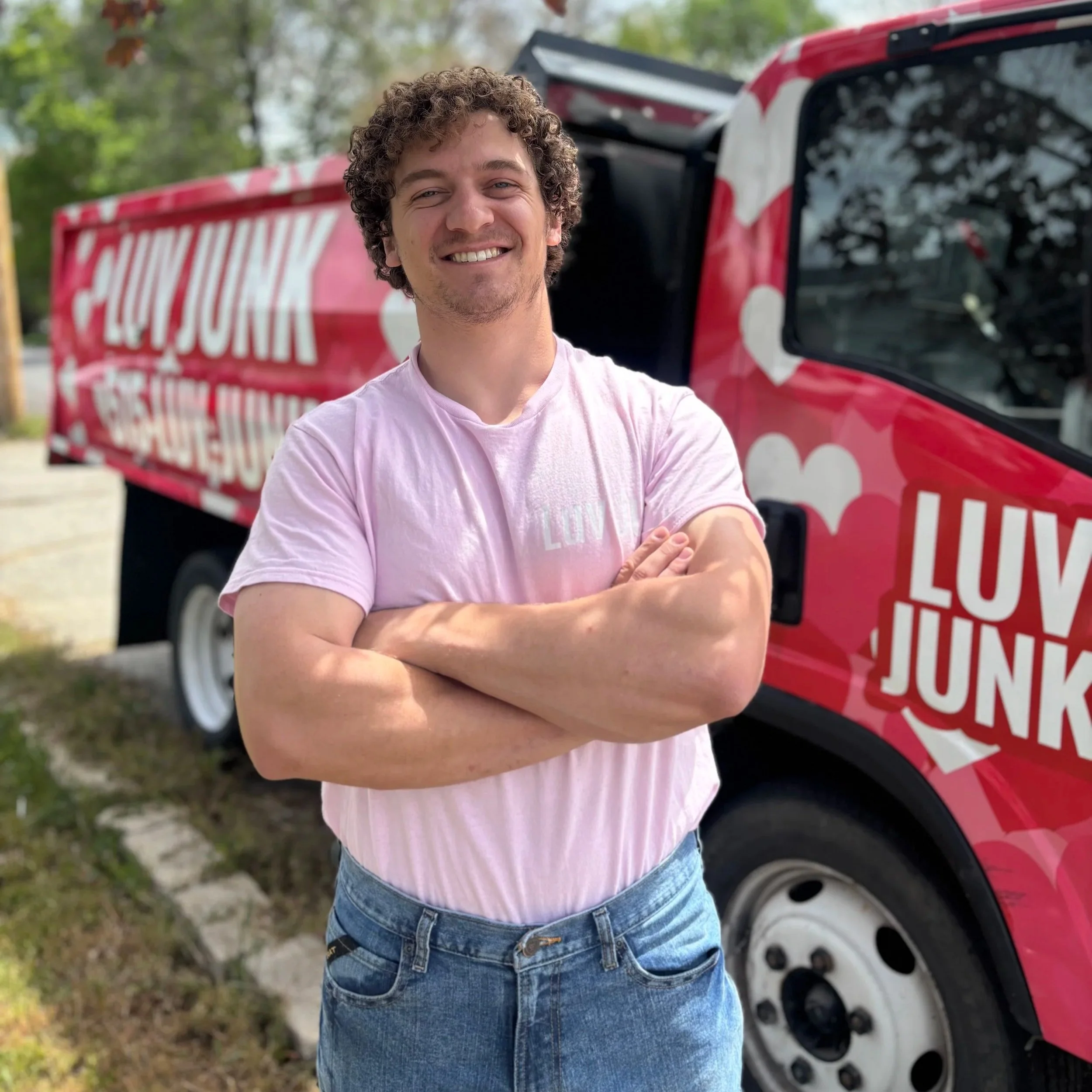 A young man with curly hair and a pink T-shirt smiling with arms crossed, standing in front of a red vehicle with white polka dots.