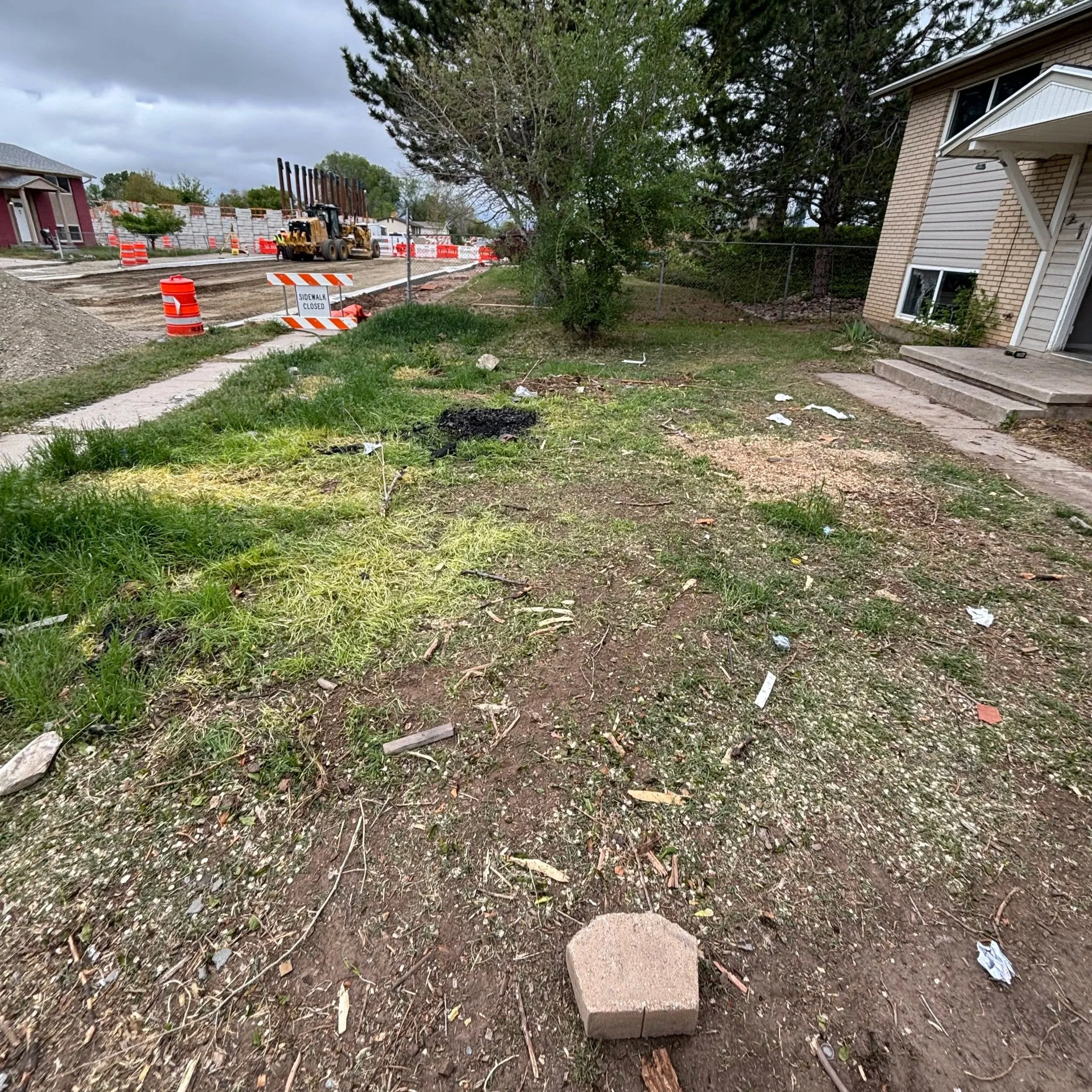 A residential street under construction with construction equipment, orange traffic barrels, a sidewalk closed sign, a damaged sidewalk, and a house on the right side. The sky is cloudy.