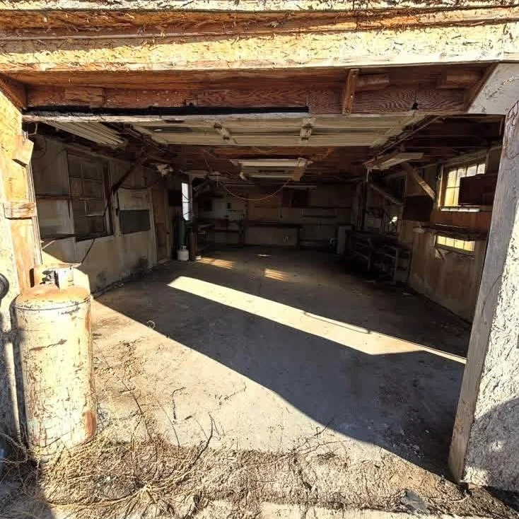 Empty garage or workshop with dirt floor, some sunlight entering through windows, and shelves on the back wall.