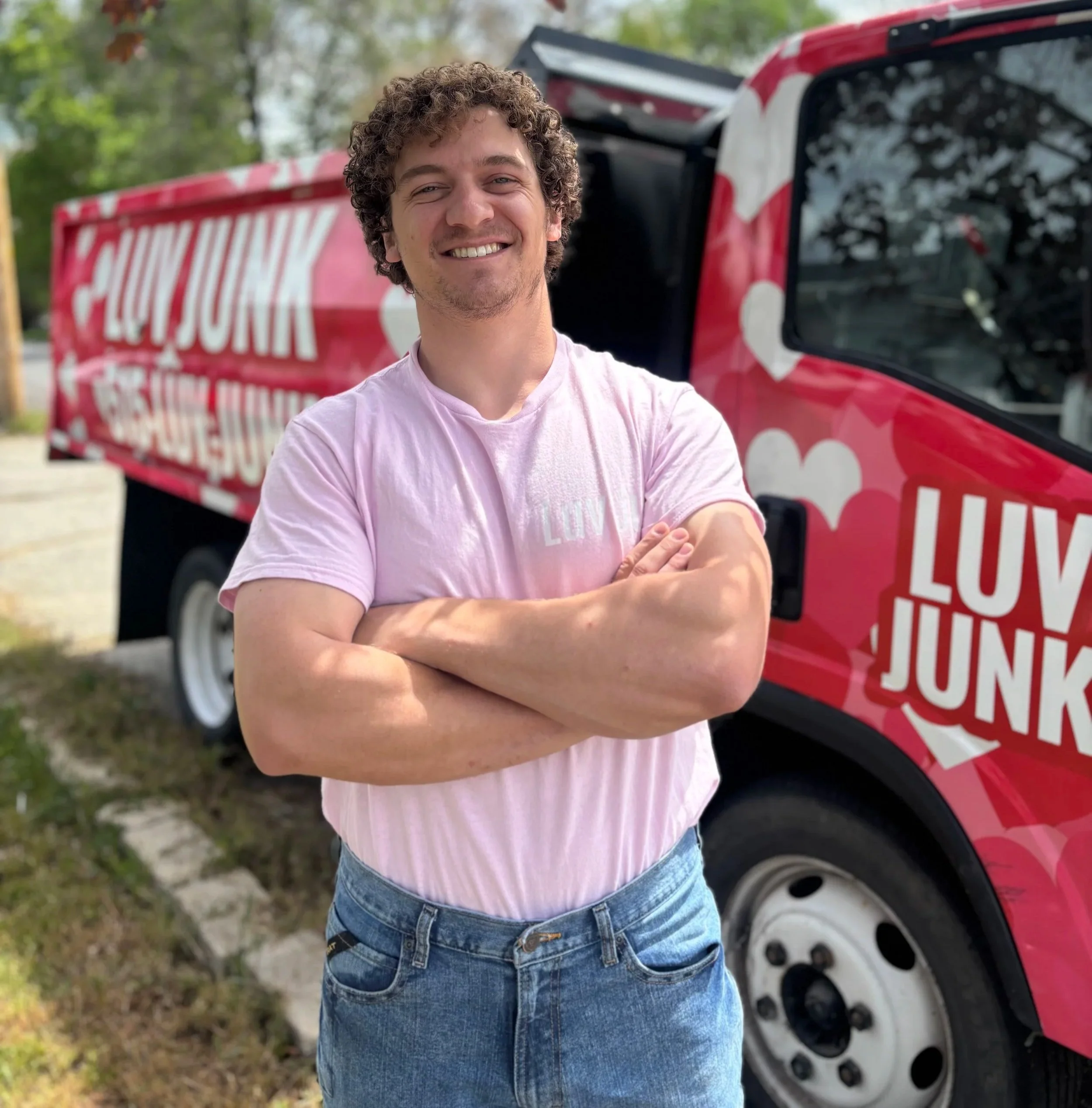 A person with curly hair smiling and crossing their arms, standing in front of a red vehicle with white polka dots and a black window, outdoors with trees in the background.