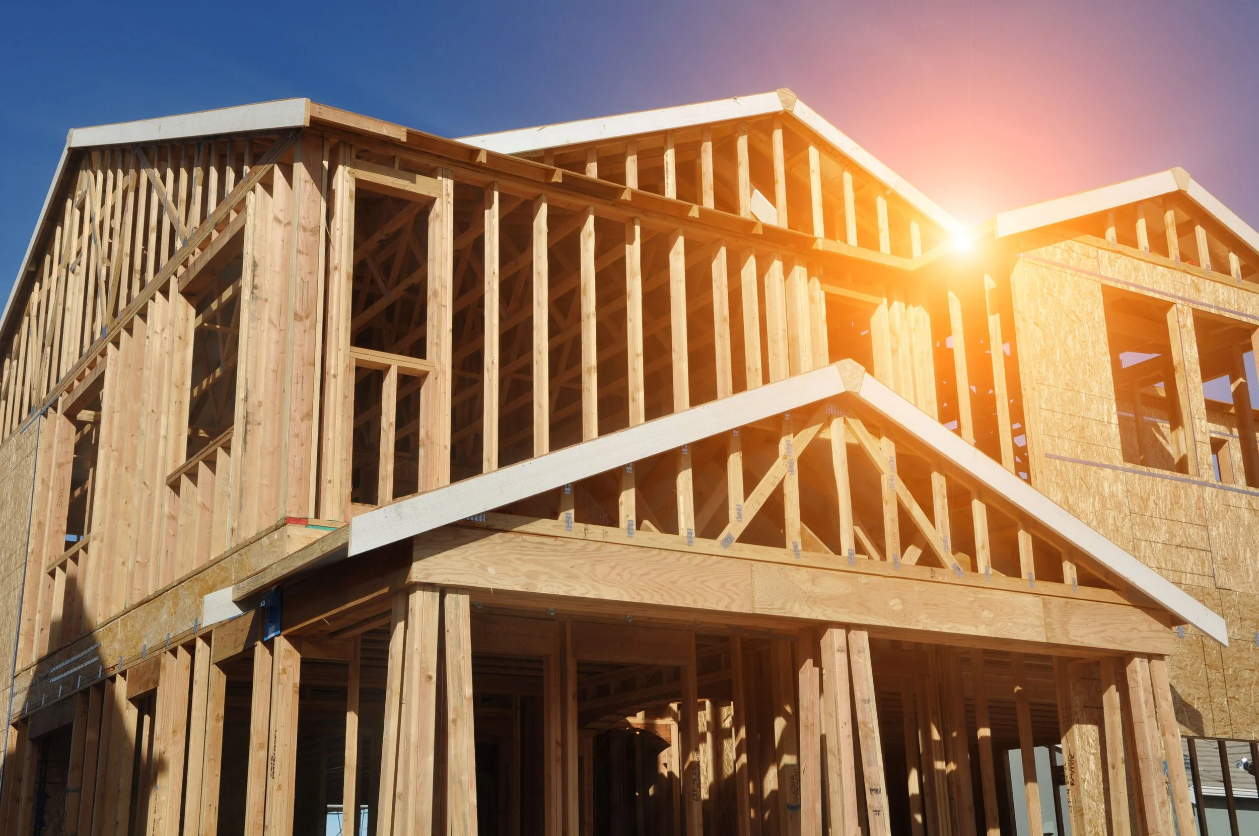 Wooden house under construction with exposed framing, set against a blue sky with sunlight shining.