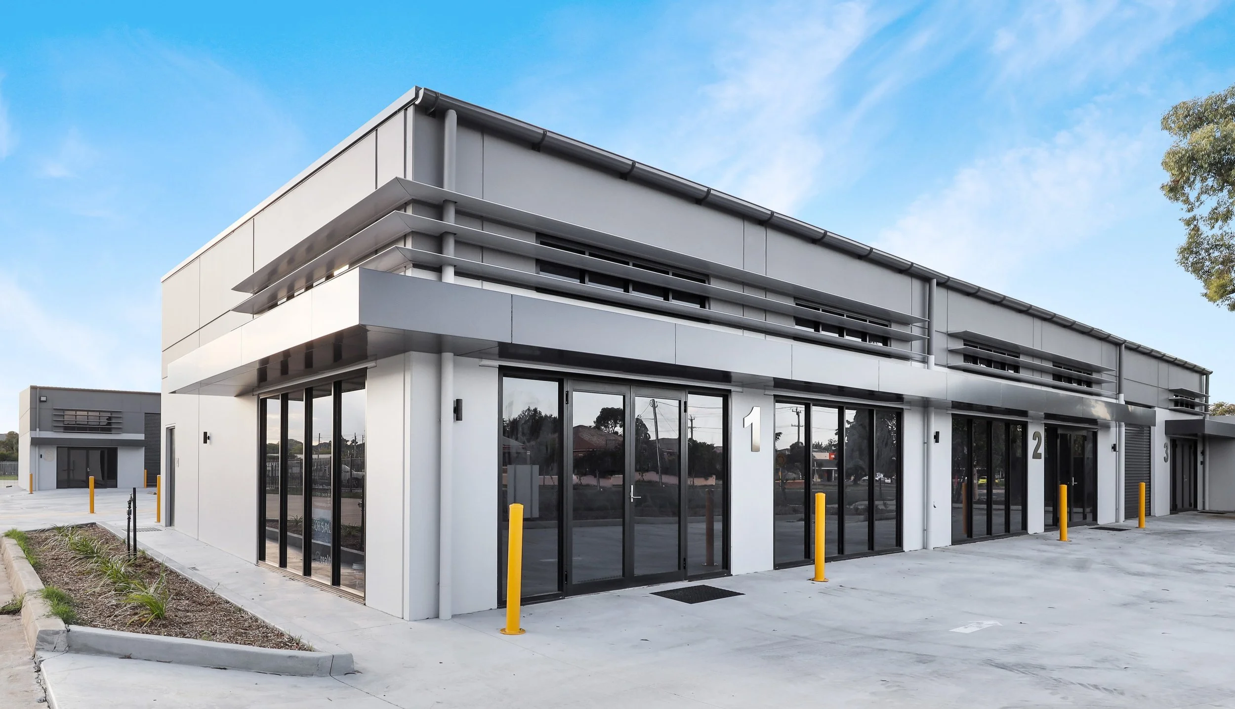 Modern commercial building with large glass doors, featuring sleek white walls, black accents, and a flat roof, set against a blue sky with some trees in the background.