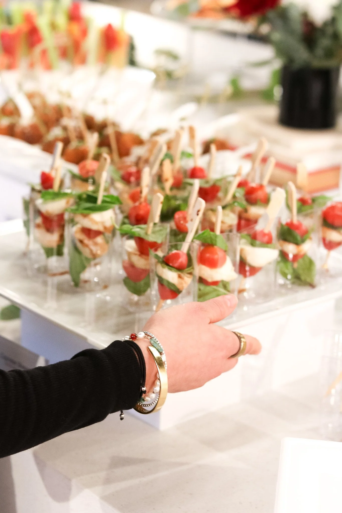 A hand reaching for a glass cup filled with a layered appetizer with tomatoes, basil, and cheese, set on a white serving tray at a party or buffet.