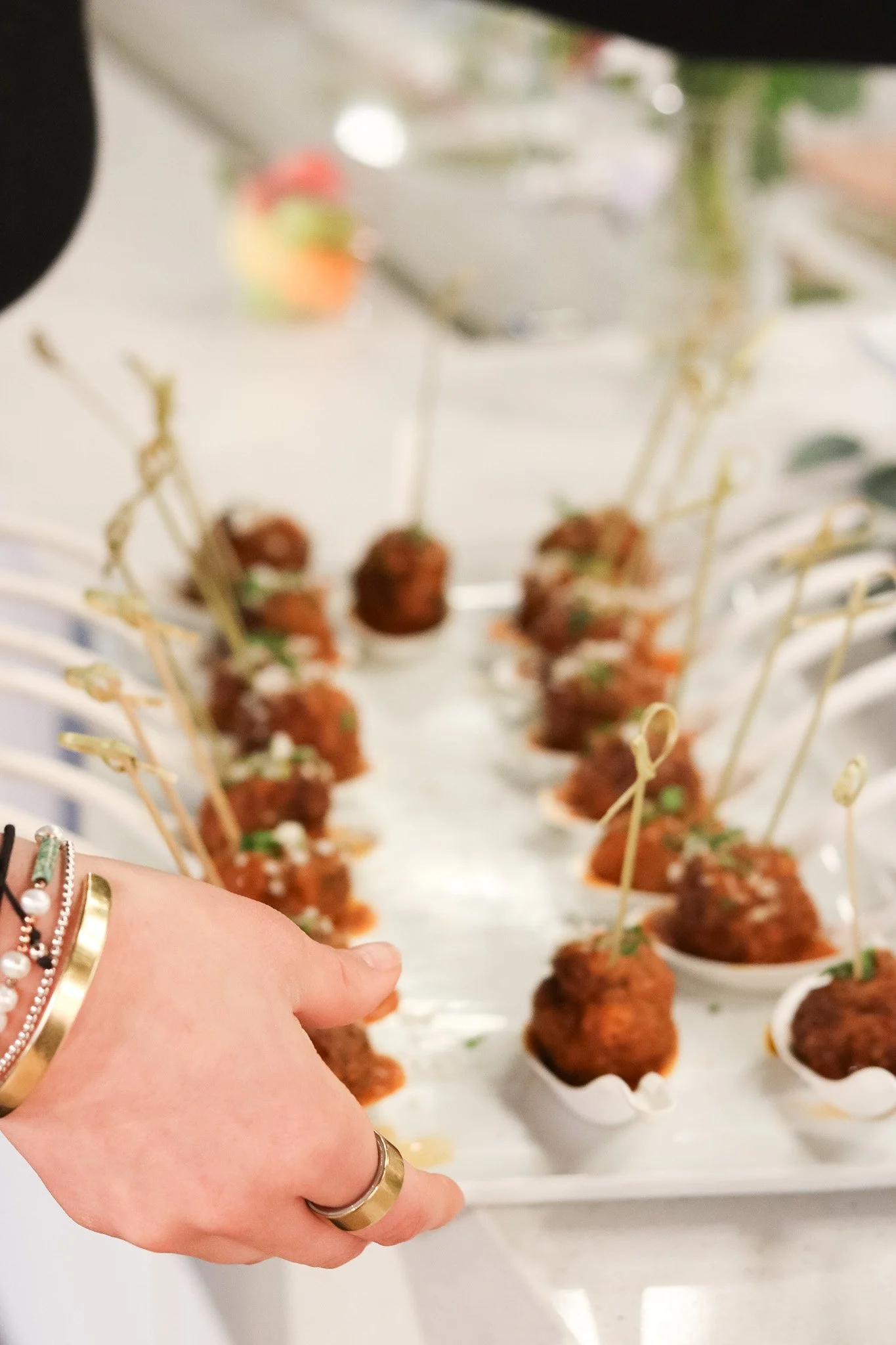 A hand reaching for a platter of cocktail meatballs served in white cups, with toothpicks, on a table at a gathering or party.