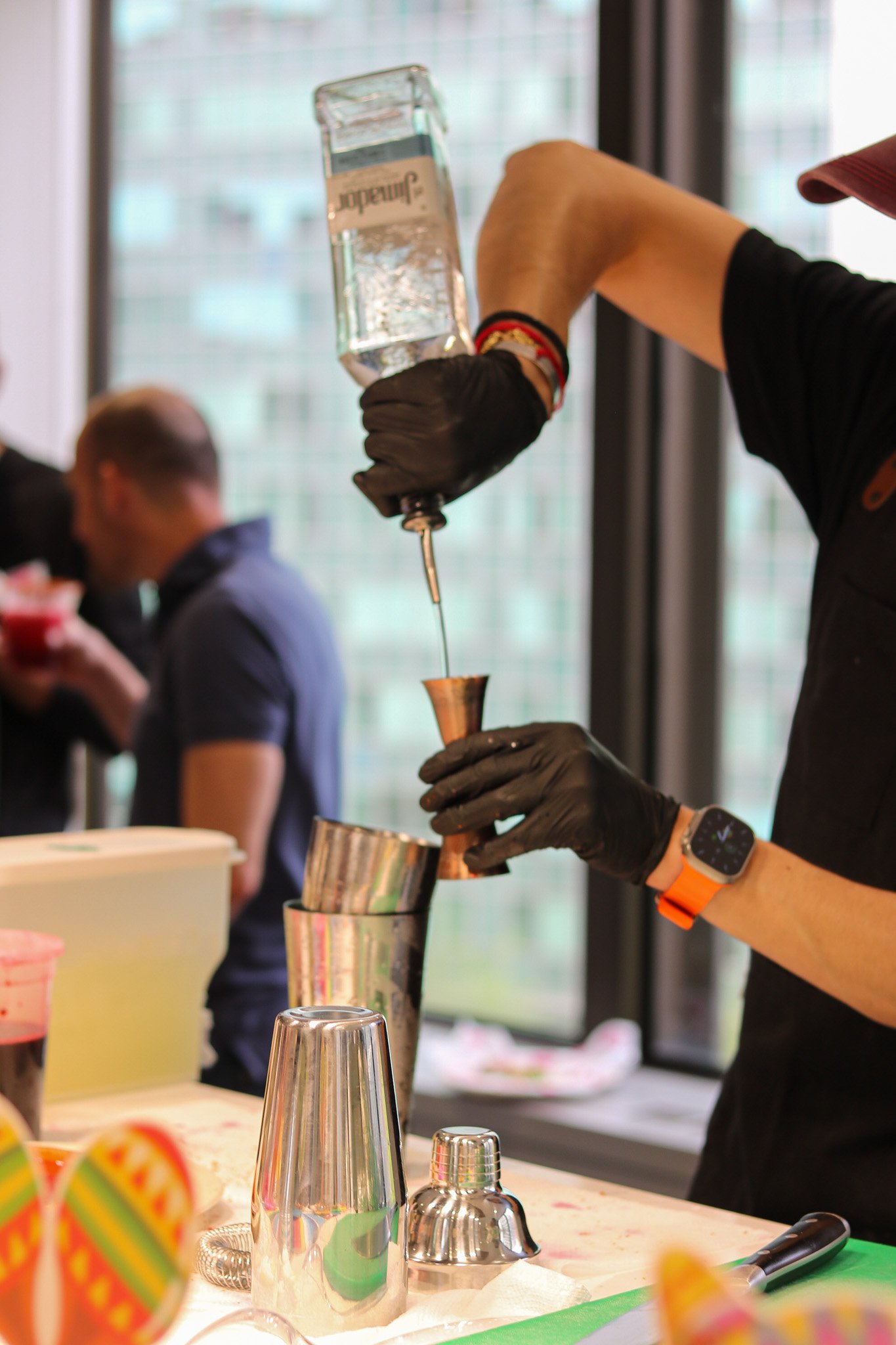 Bartender pouring liquid into a metal jigger at a bar with bar tools and colorful decorations in the background.