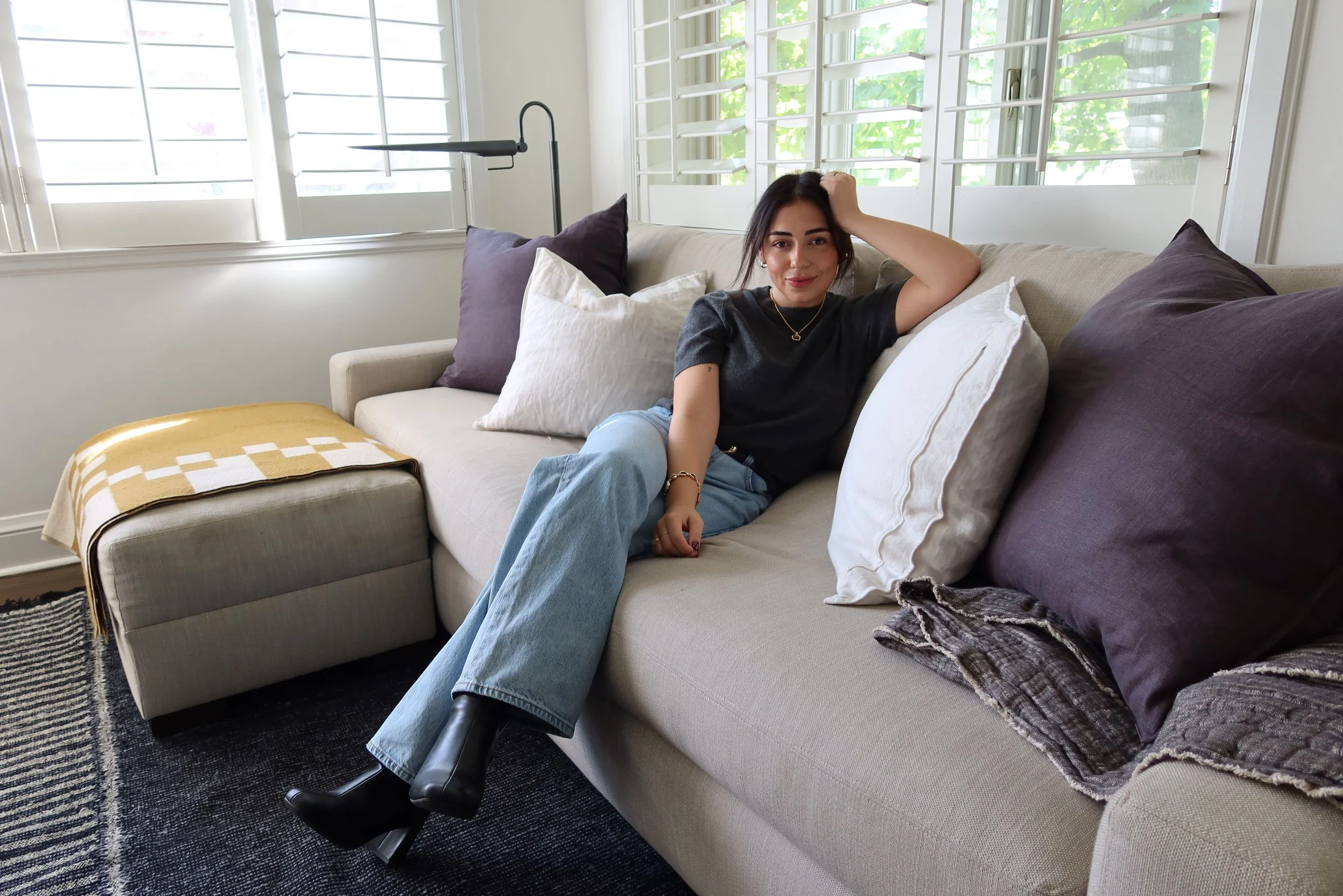 A woman sitting on a beige sofa in a bright room with large windows and white shutters. She is wearing a black T-shirt, light blue jeans, and black boots, with a gold necklace and bracelet. She is smiling, with styled dark hair, and her right arm resting on a white pillow while her left hand touching her head.