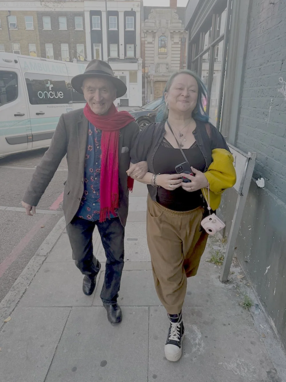 Martin Carthy and Eliza Carthy walking arm in arm down a London street after the first day of rehearsals in New Cross. Martin wears a wide-brimmed hat, patterned shirt and red scarf, while Eliza smiles beside him, holding her phone and camera.