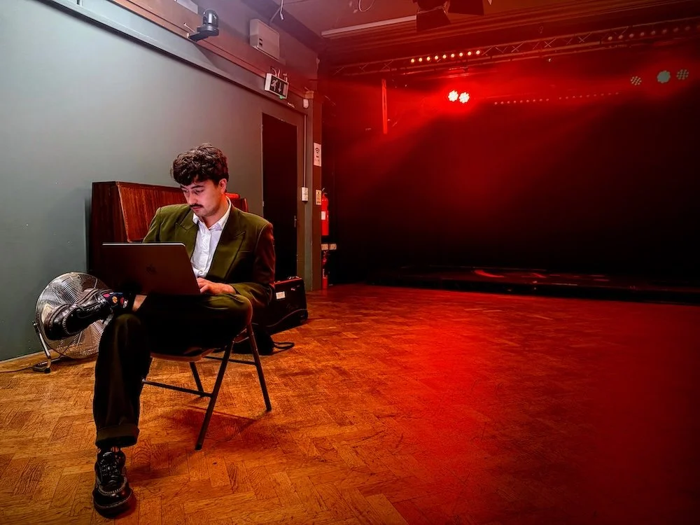 Campbell Baum of Broadside Hacks sits on a chair backstage, working on a laptop, lit by red stage lights that cast a glow across the empty floor and black backdrop.