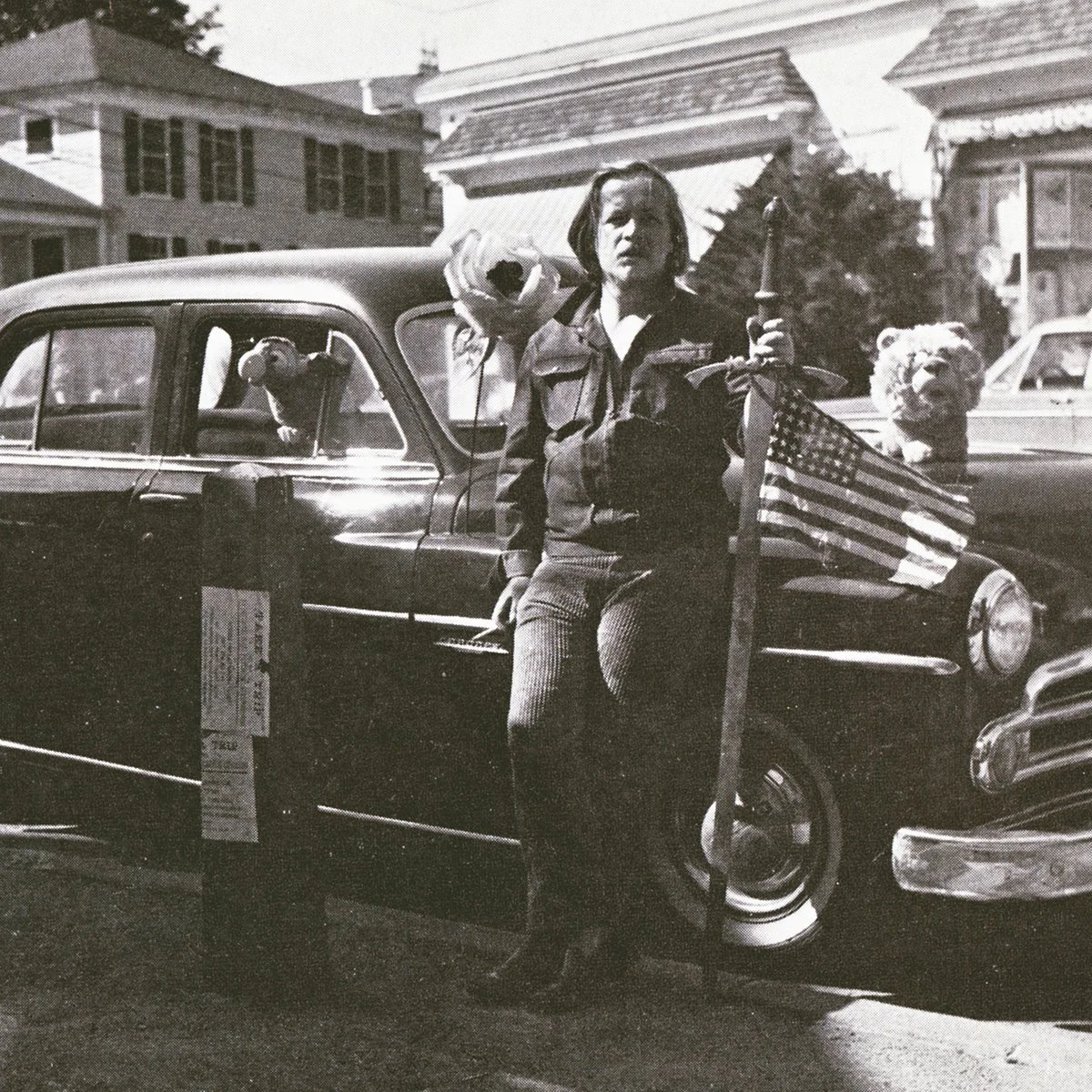 Jackson C. Frank in Buffalo, mid-1960s, standing beside a car holding a ceremonial sword with a US flag attached to the hilt, with a dog perched on the car behind him.