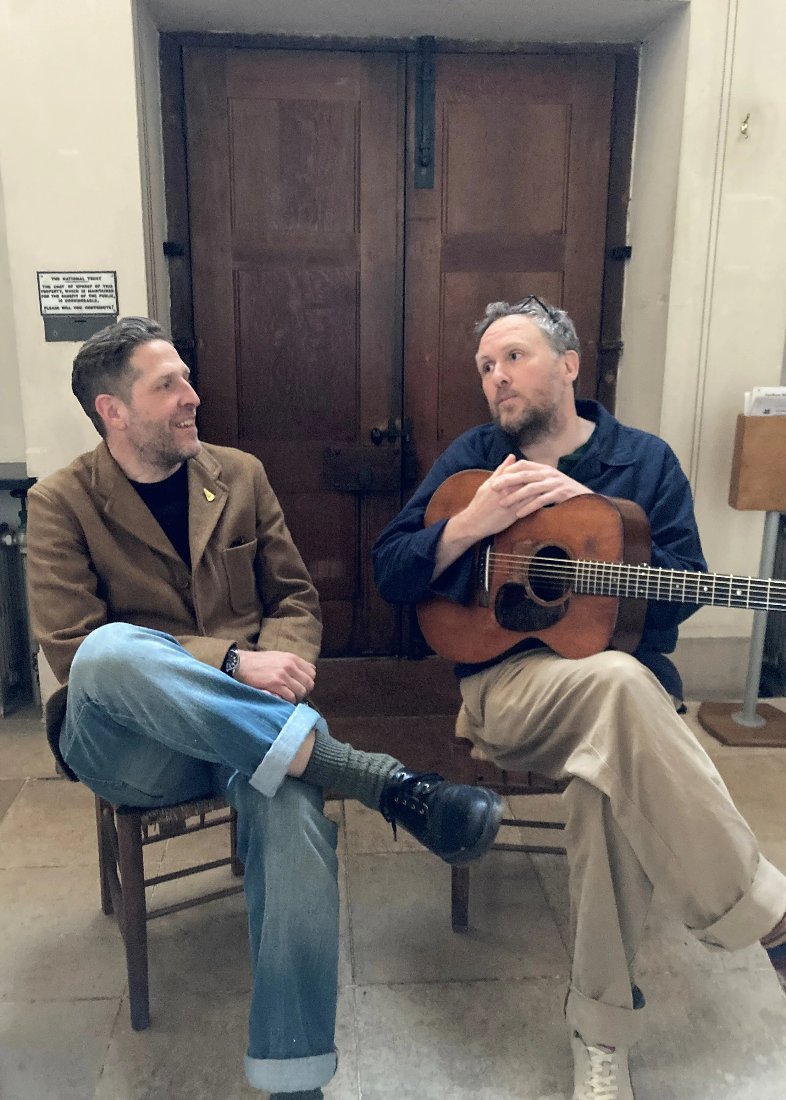 Richard Lines and Jon Wilks seated inside Sandham Memorial Chapel, mid-conversation. Jon holds an acoustic guitar; a National Trust notice is visible on the wall behind them.