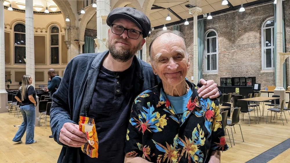 Jon Wilks and Martin Carthy smiling together at Manchester Folk Festival, standing in a hall with tall white columns, wooden floor, and scattered chairs in the background.