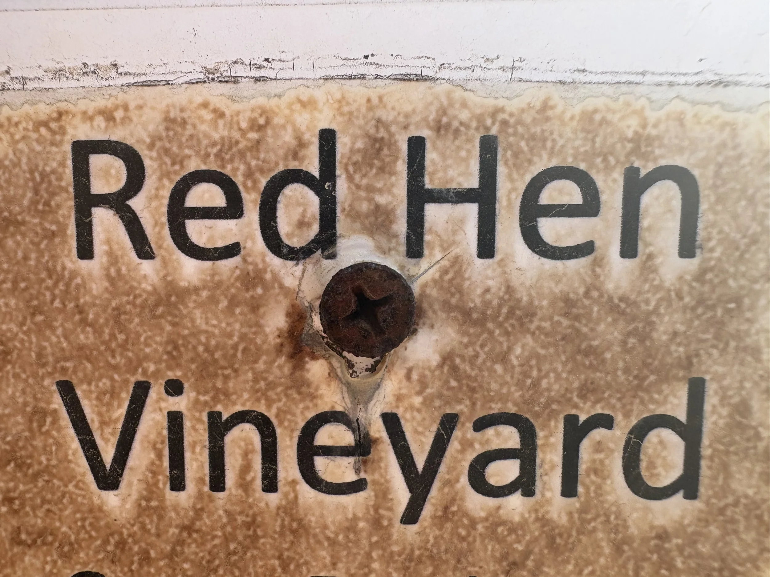 A metal plate with the words "Red Hen Vineyard" printed on it, attached to a wall with a screw in the center.