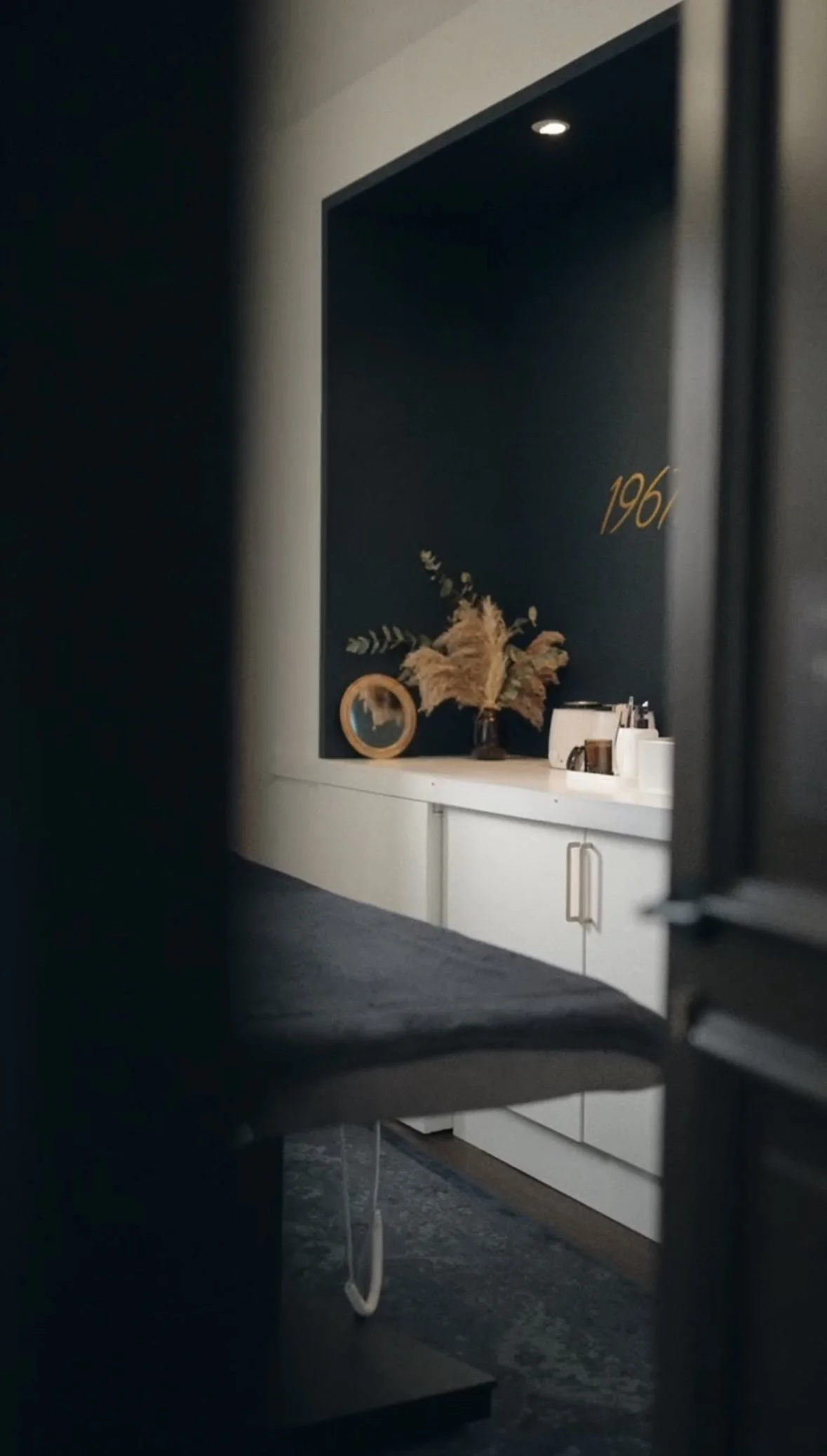 Un comptoir blanc avec des décorations, dont un vase avec des plantes séchées, un miroir rond et quelques objets. Le mur derrière est bleu foncé avec une inscription dorée '1964'.